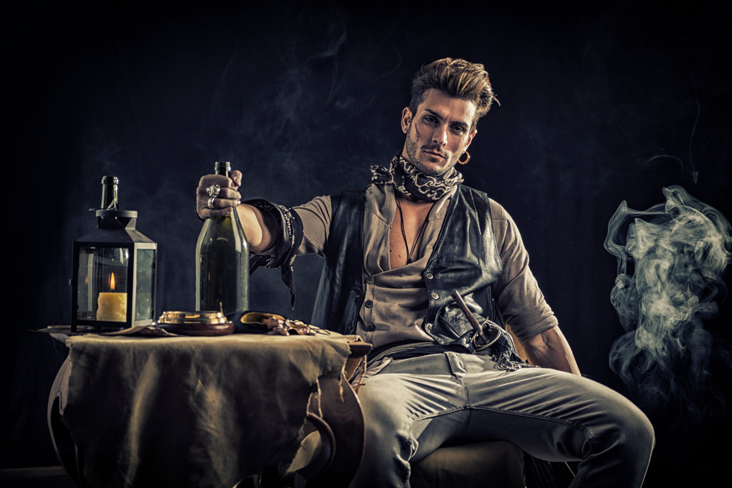 A young man with styled hair, wearing a scarf, earrings, and casual clothing, sits at a table with bottles, a lantern, and food, surrounded by smoke and dark background.