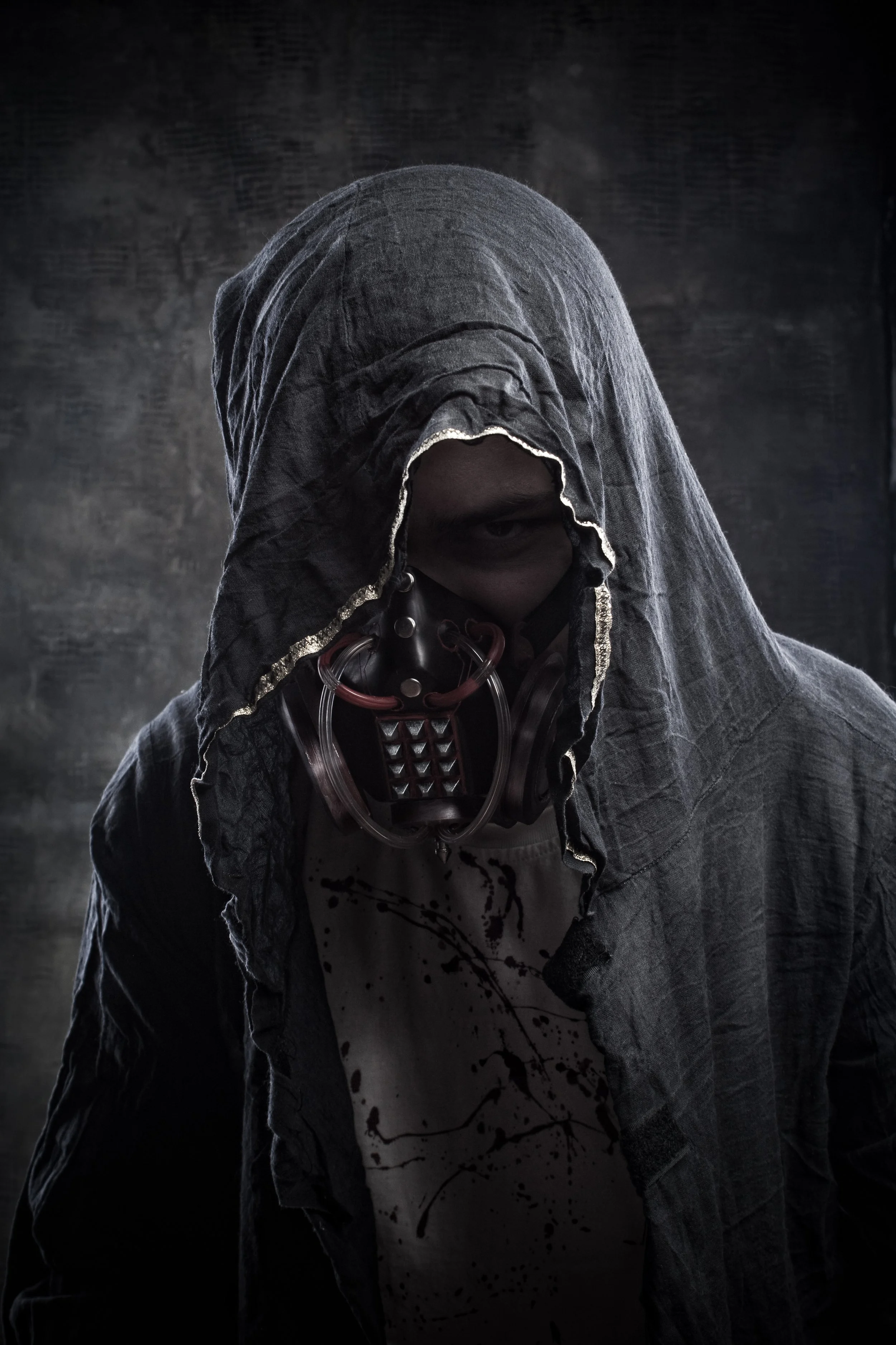 A person wearing a dark hooded cloak with a textured fabric, covering most of their face. Their face is partially visible with dark eye makeup, and they are wearing a mask around their neck with metallic and leather details. The background is dark and slightly textured.