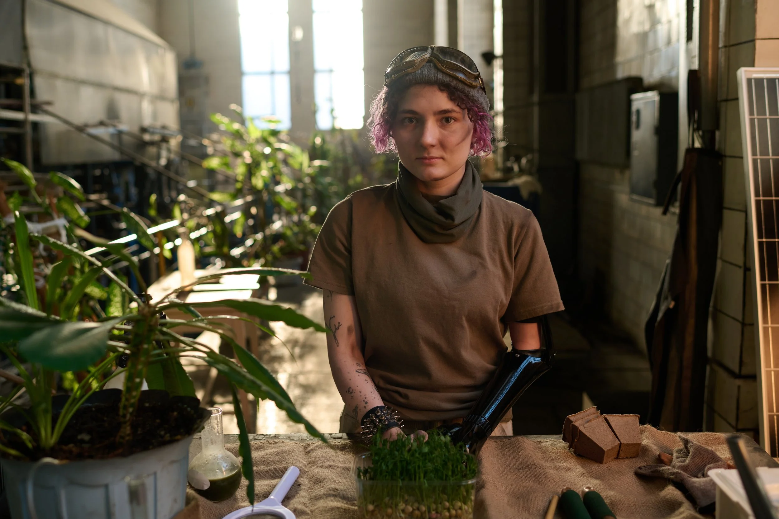 A young person with pink hair, wearing goggles on their head, working in a greenhouse with plants and gardening tools.