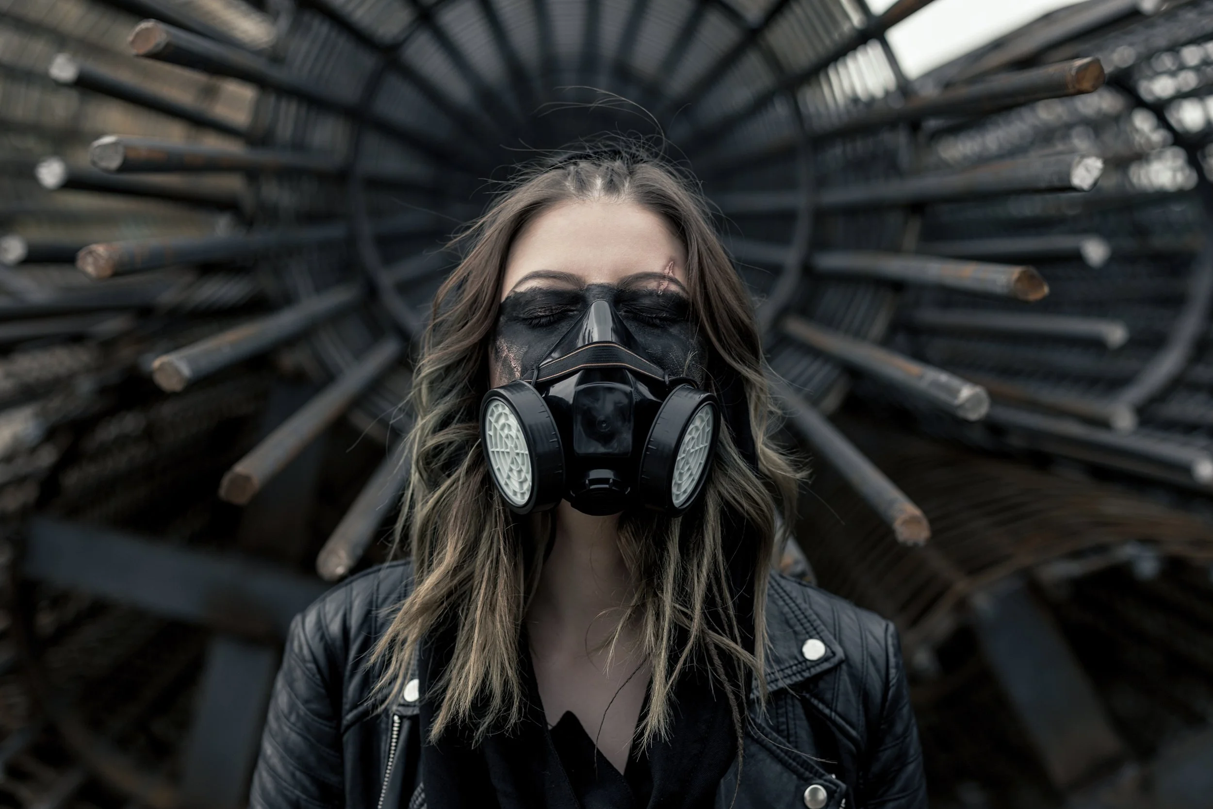A woman wearing a black gas mask and a black leather jacket, with long wavy hair, stands in front of a circular structure made of metal pipes.