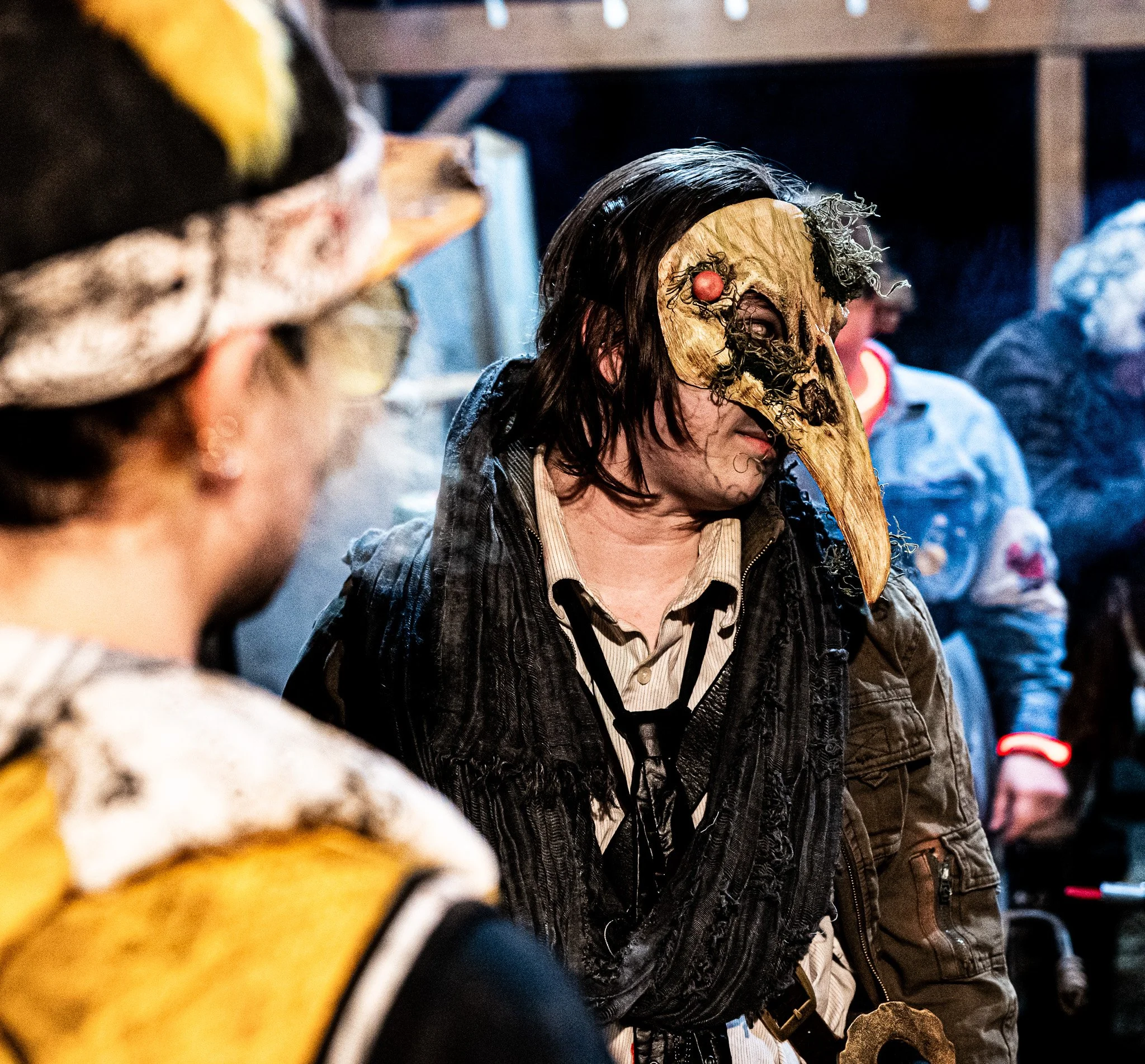 A person wearing a mask designed as a long beak, resembling a plague doctor mask, with strands of hair around the face, standing in a row with other people also wearing masks, in a dimly lit environment.