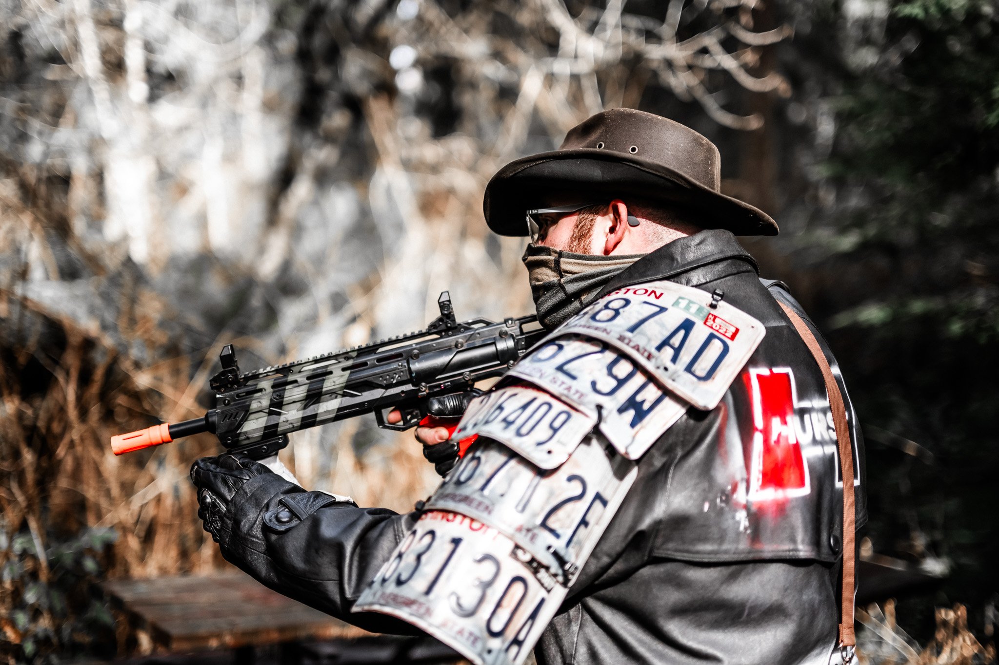 A person wearing a wide-brimmed hat, glasses, and a face covering holding a rifle in a wooded outdoor setting.