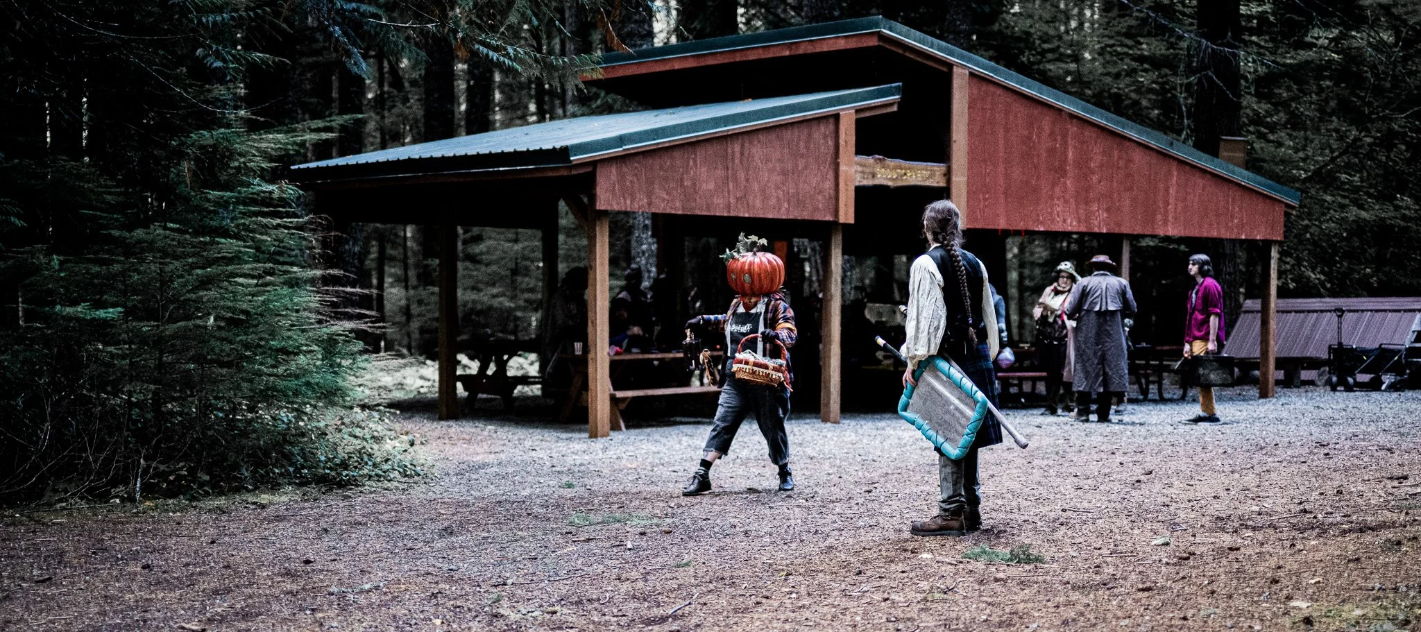People gathered at an outdoor forested area with a red wooden shelter, some wearing costumes, with one person dressed as a pumpkin, during daytime.