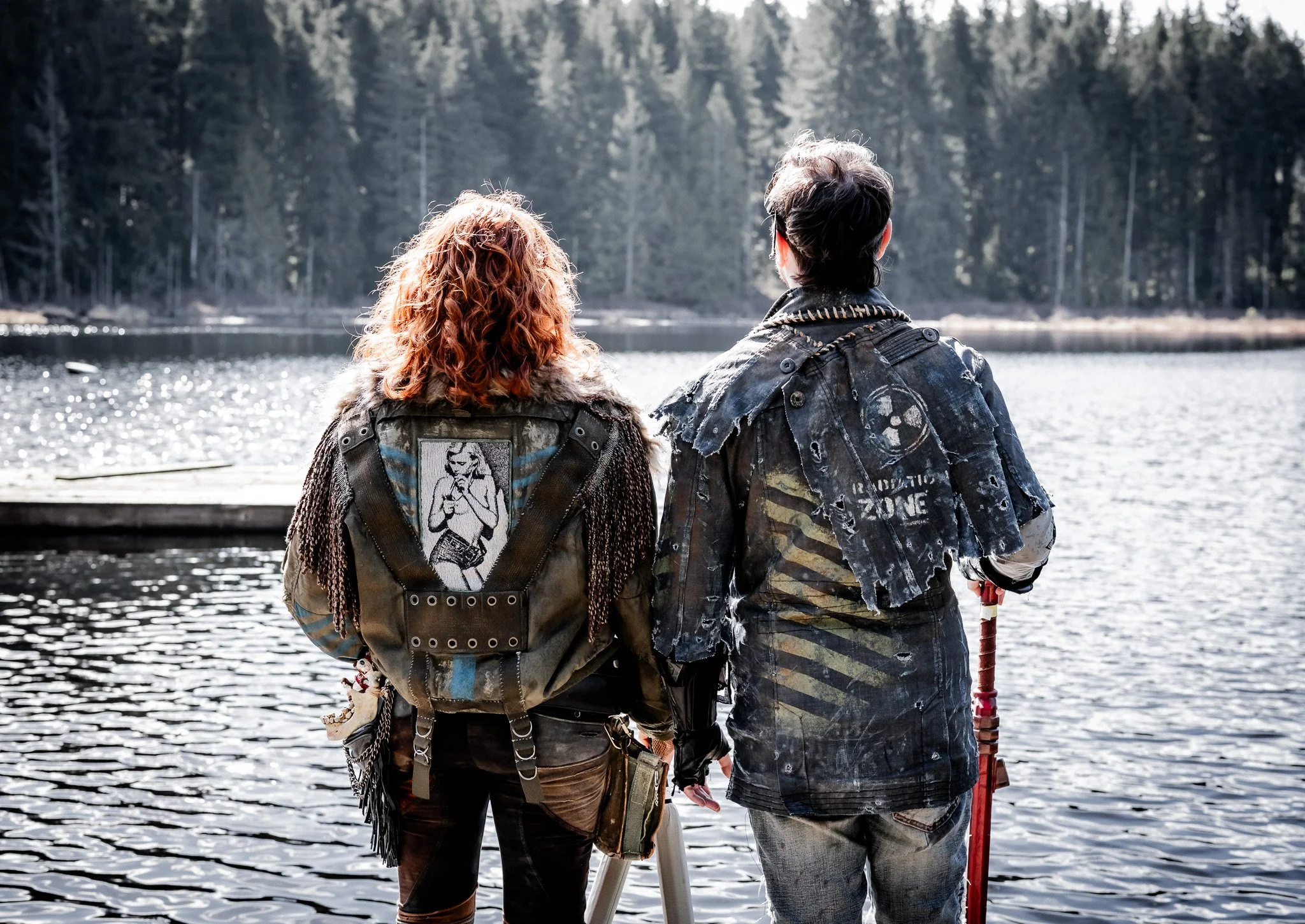 Two people with back view standing by a lake, surrounded by a dense forest, during daylight.