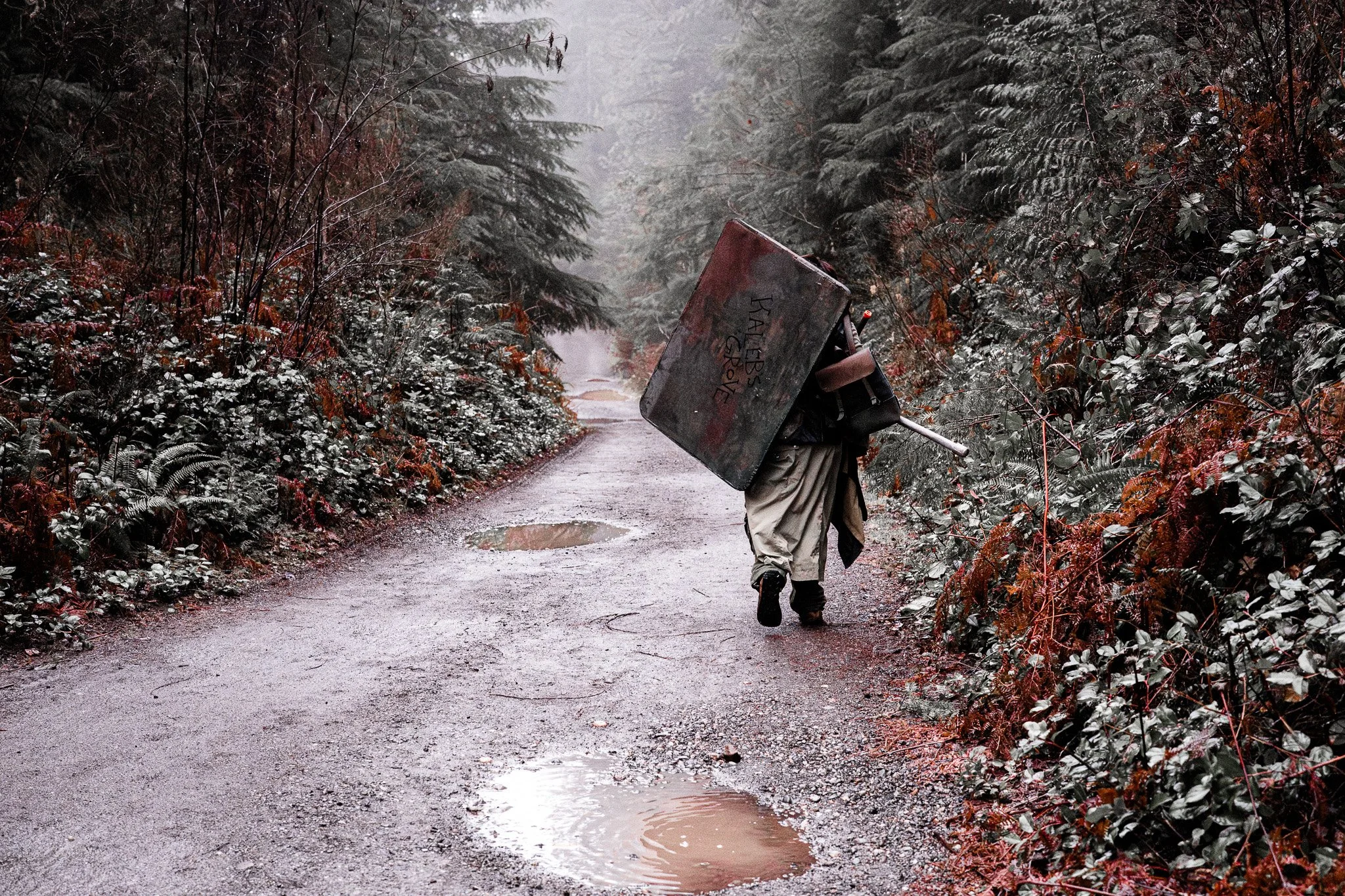 A person walking down a dirt road in a forest, carrying a large, rusted metal box labeled 'Kalabis Grave' on their back, with overgrown plants and trees surrounding the scene.