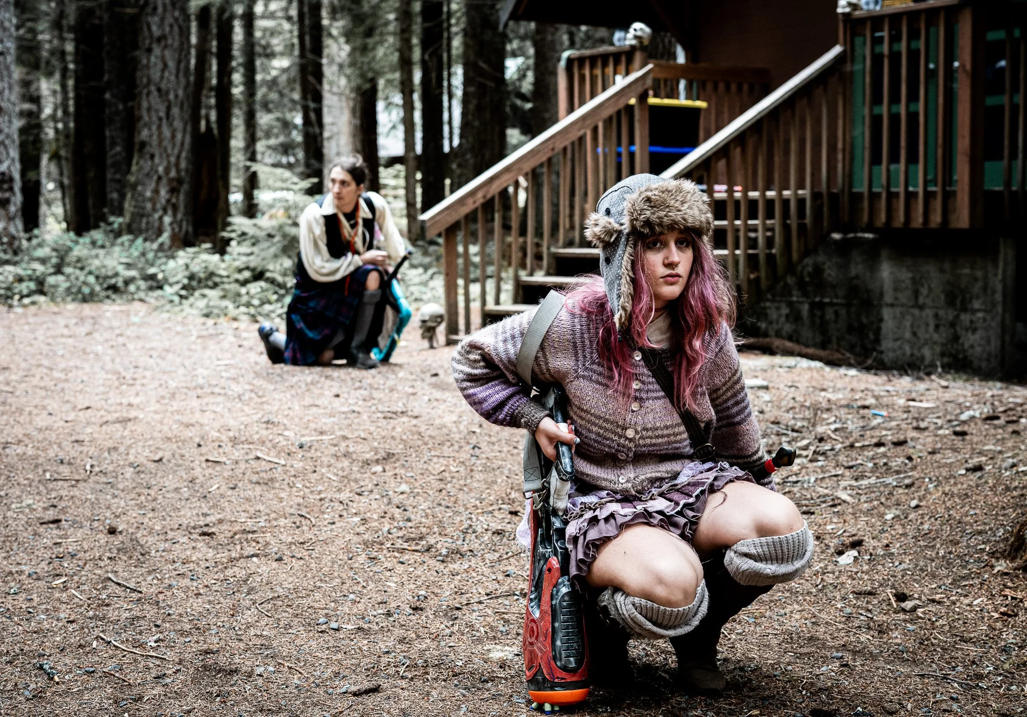A young woman with pink hair in a hat and sweater kneeling on the ground, while another person with dark hair and a beige coat kneels behind her in a wooded outdoor setting near a wooden house.