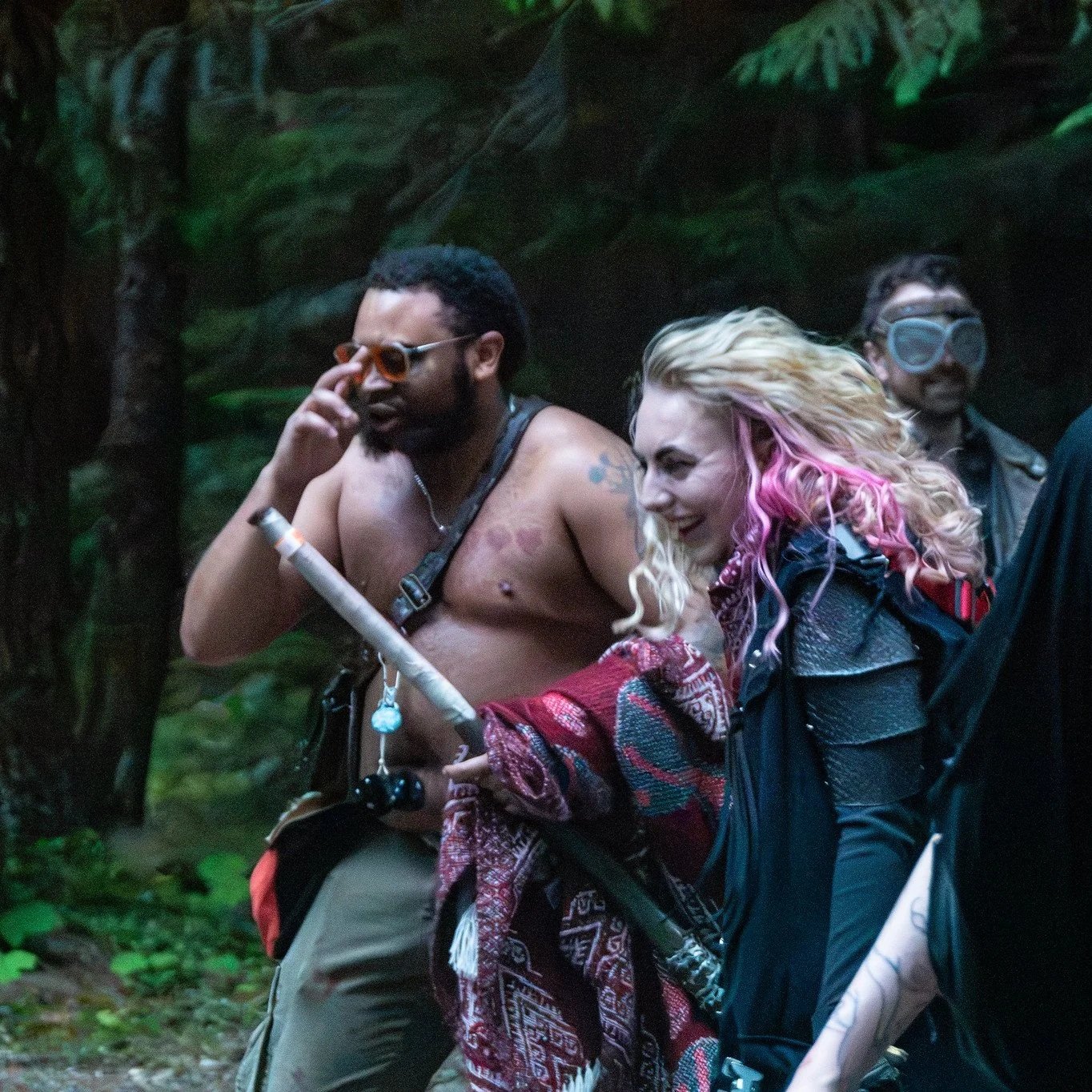 Three people walking in a lush, green forest, one shirtless man with tattoos and sunglasses, a woman with multicolored curly hair and a black jacket, and another man wearing glasses and a dark jacket.
