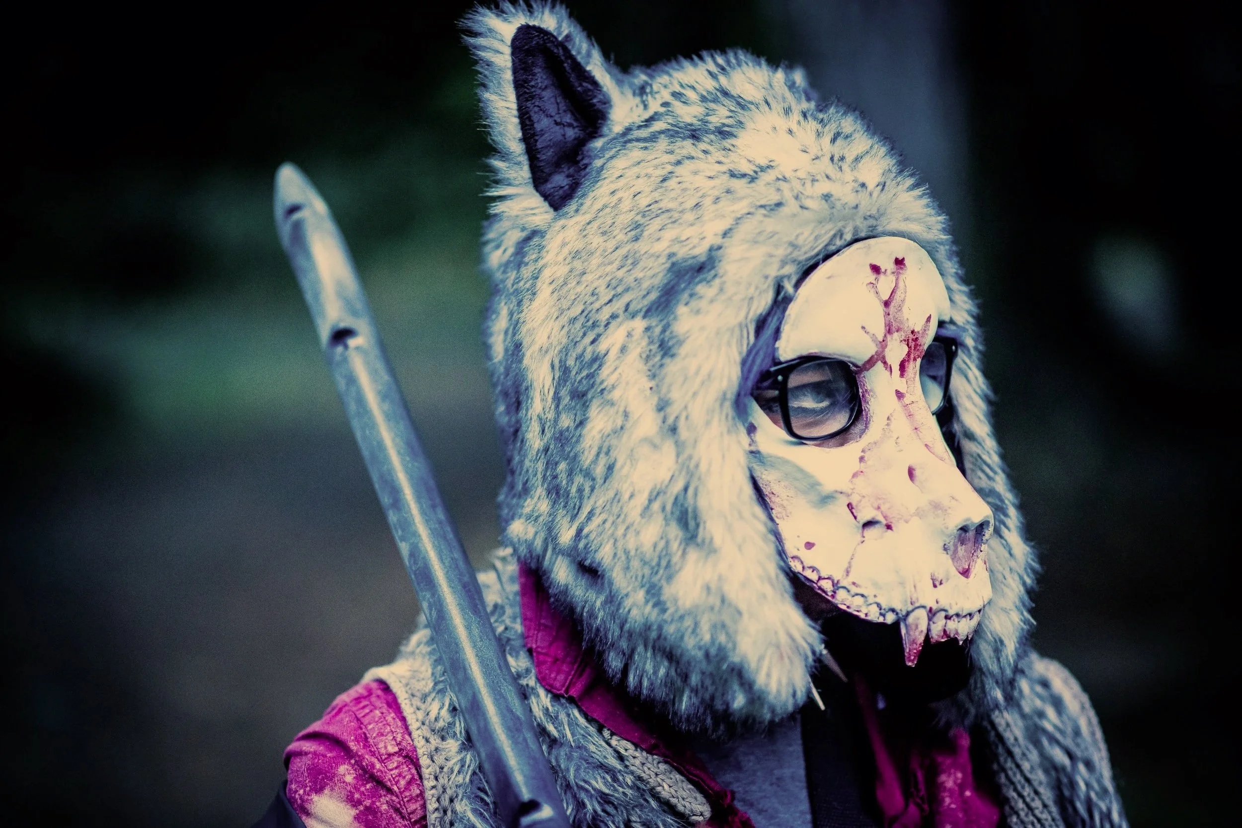 A person wearing a wolf mask with glasses and a pink jacket, holding a spear against a dark background.