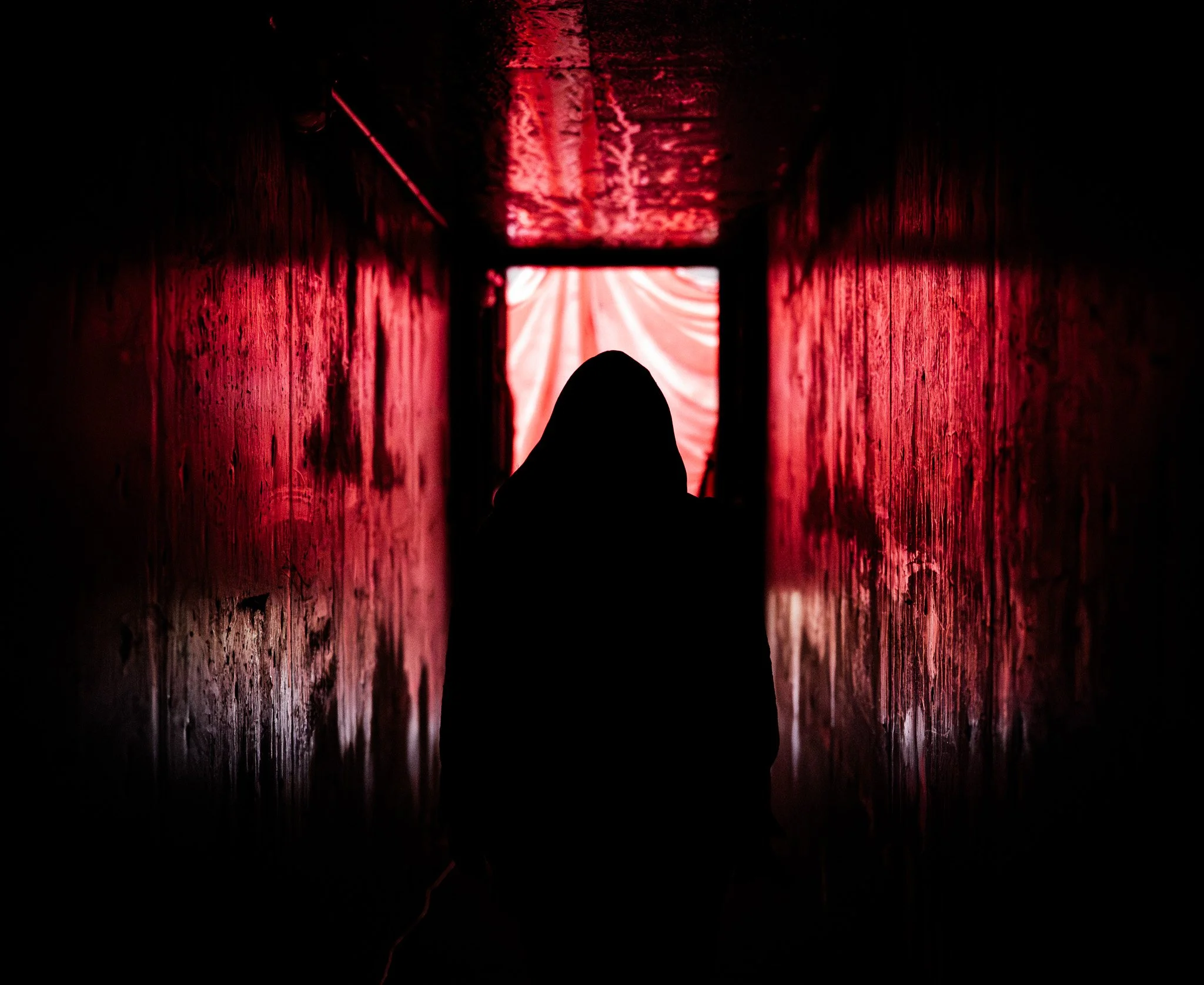 A person walking down a dark, narrow, brick-walled corridor with red lighting, towards an illuminated doorway at the end covered with fabric.