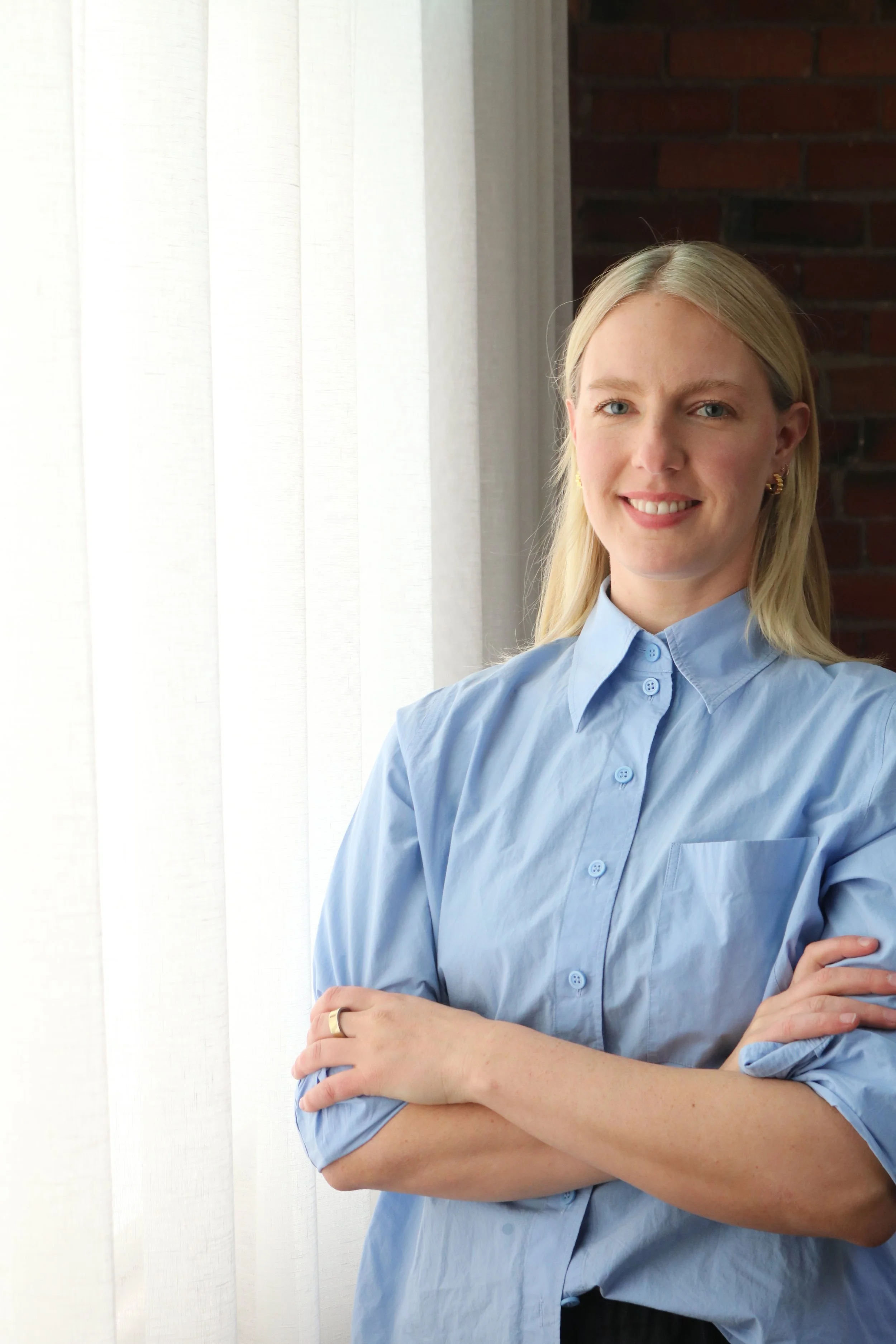 A woman with blonde hair, wearing a light blue button-up shirt, standing with arms crossed, smiling, in front of a white curtain and brick wall.