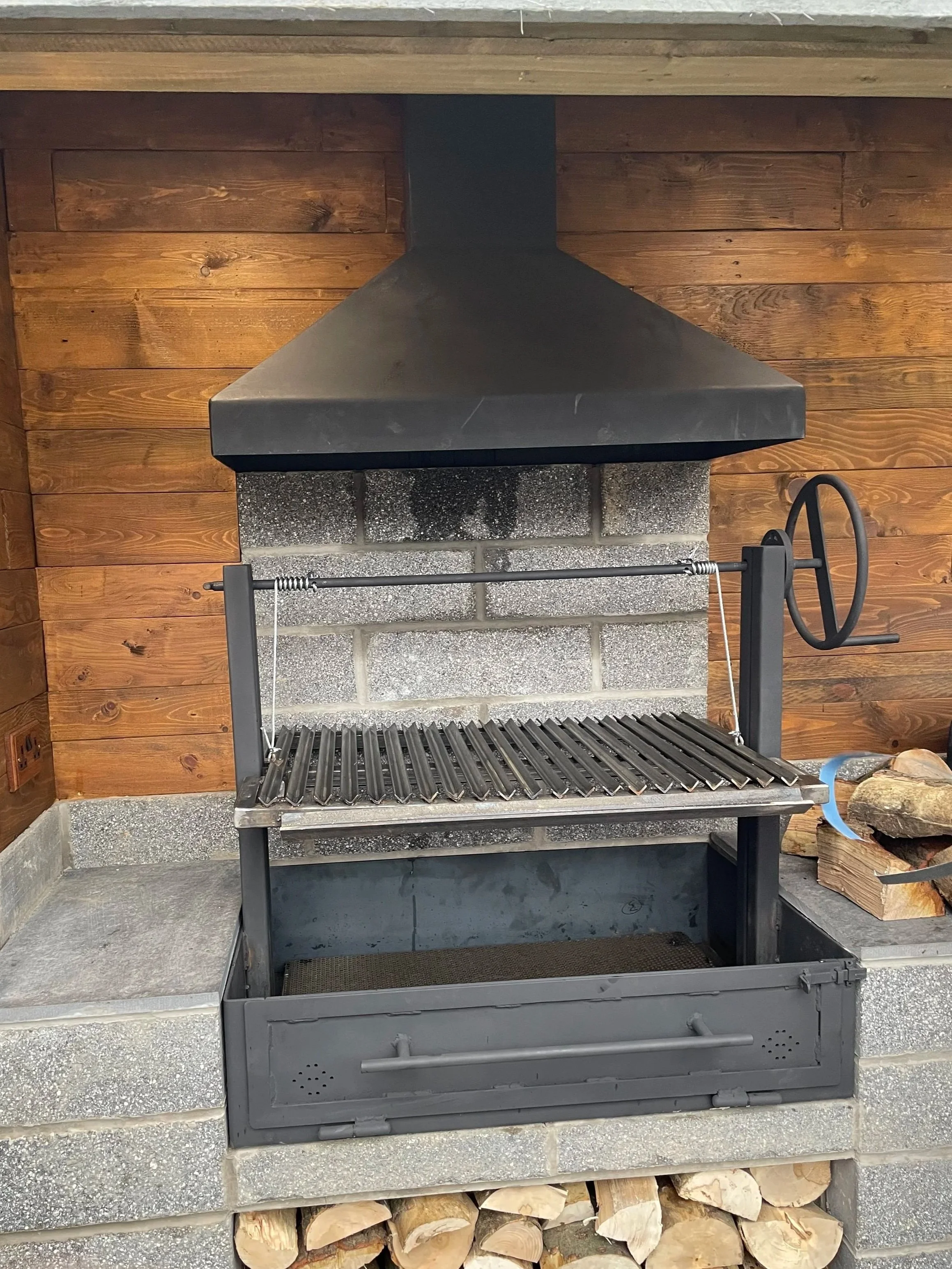 Bespoke Argentinian-style BBQ with a handwheel-adjustable cooking grill and matching steel extraction hood, made for an outdoor cooking area.