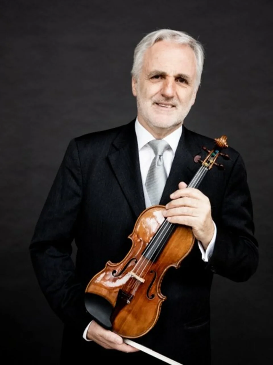 A man wearing a black suit and silver tie holding a violin against a dark background.