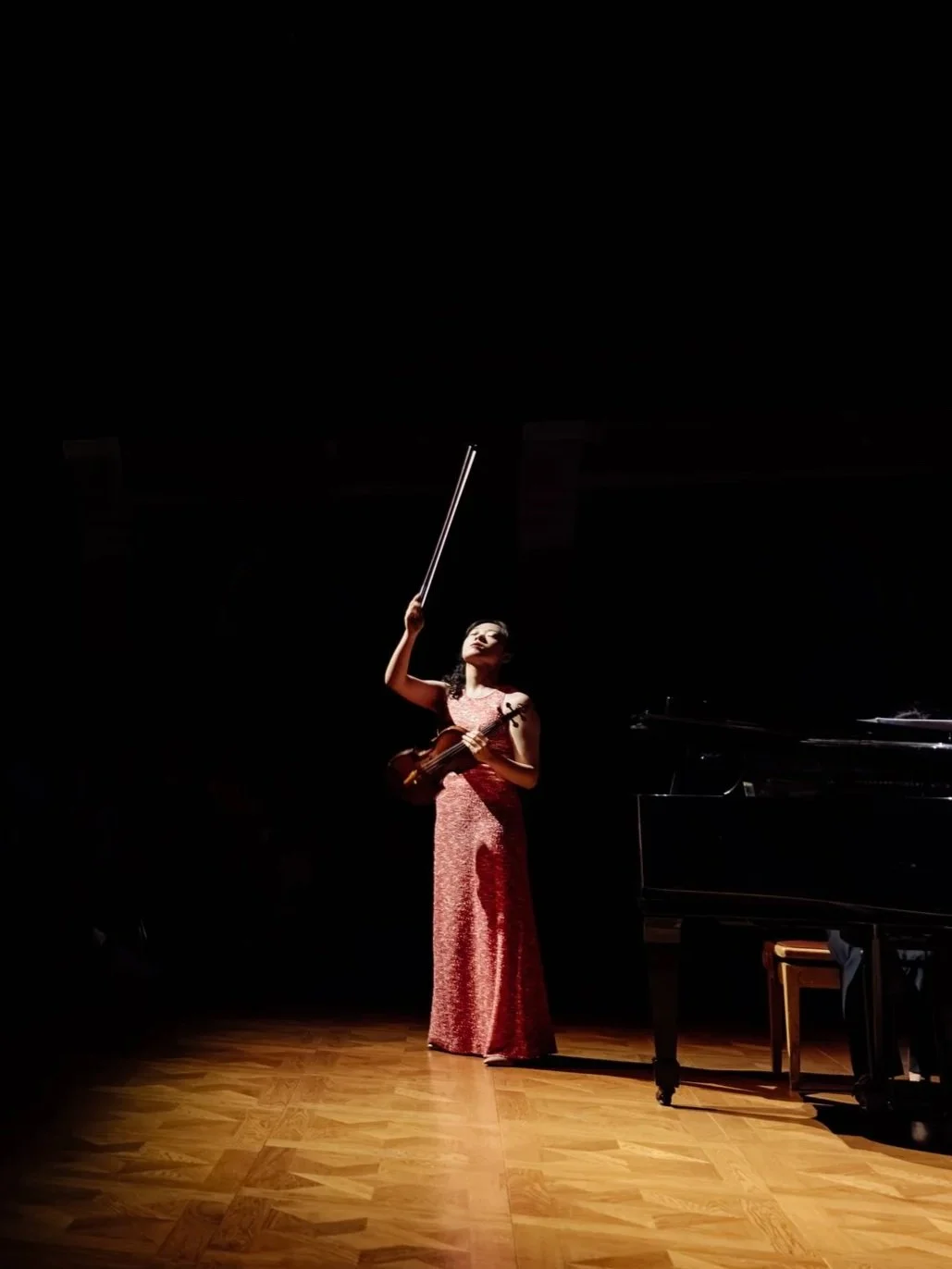A woman in a long red dress holding a violin and a bow stands on a spotlighted stage with a grand piano nearby.
