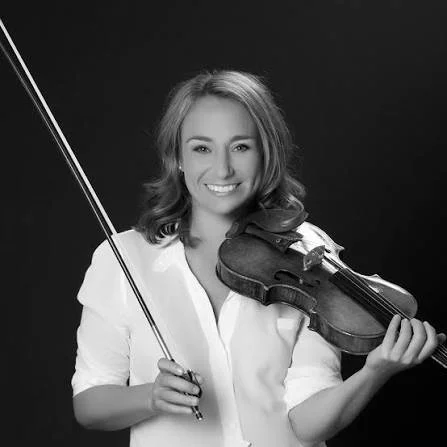 A woman smiling, holding a violin in one hand and a bow in the other.