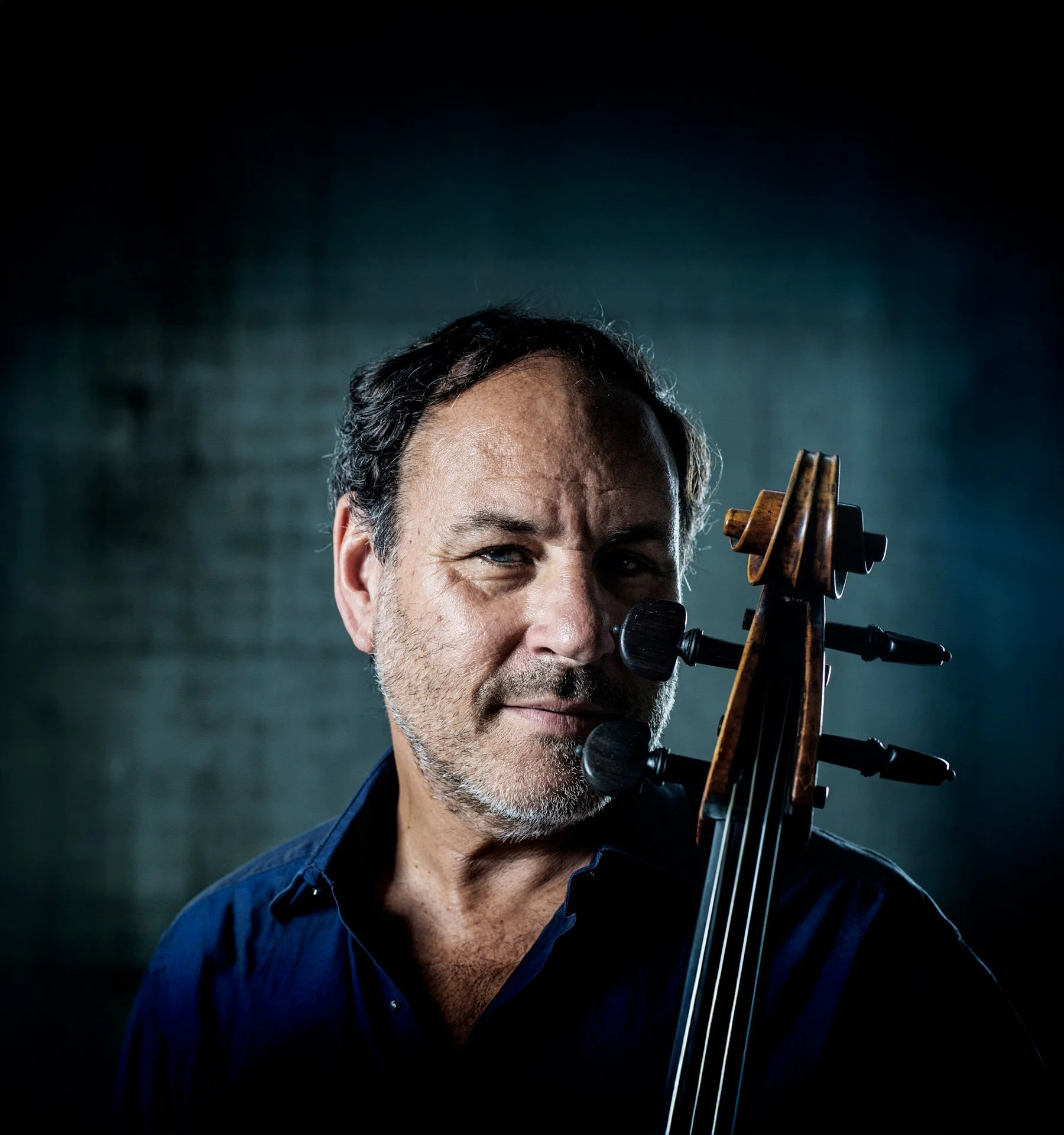 Close-up of a man with gray hair and stubble playing a cello, with the bow resting on his face against a dark background.