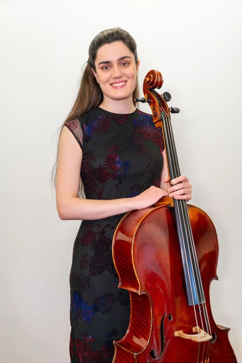 Young woman with long brown hair smiling, holding a wooden cello, standing against a plain white wall.