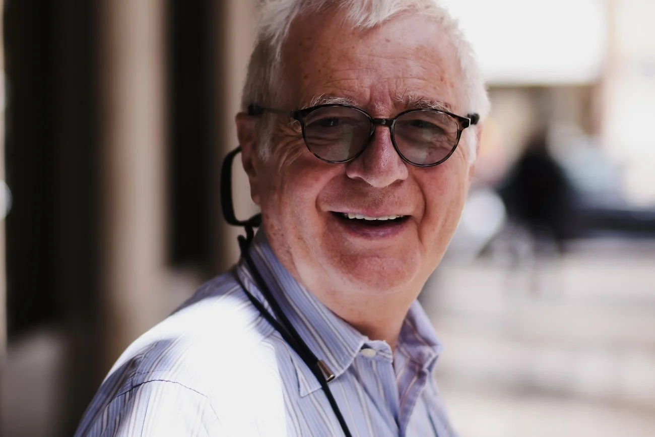 Smiling elderly man with glasses and a stethoscope around his neck, indoors or outdoors, in a professional setting.