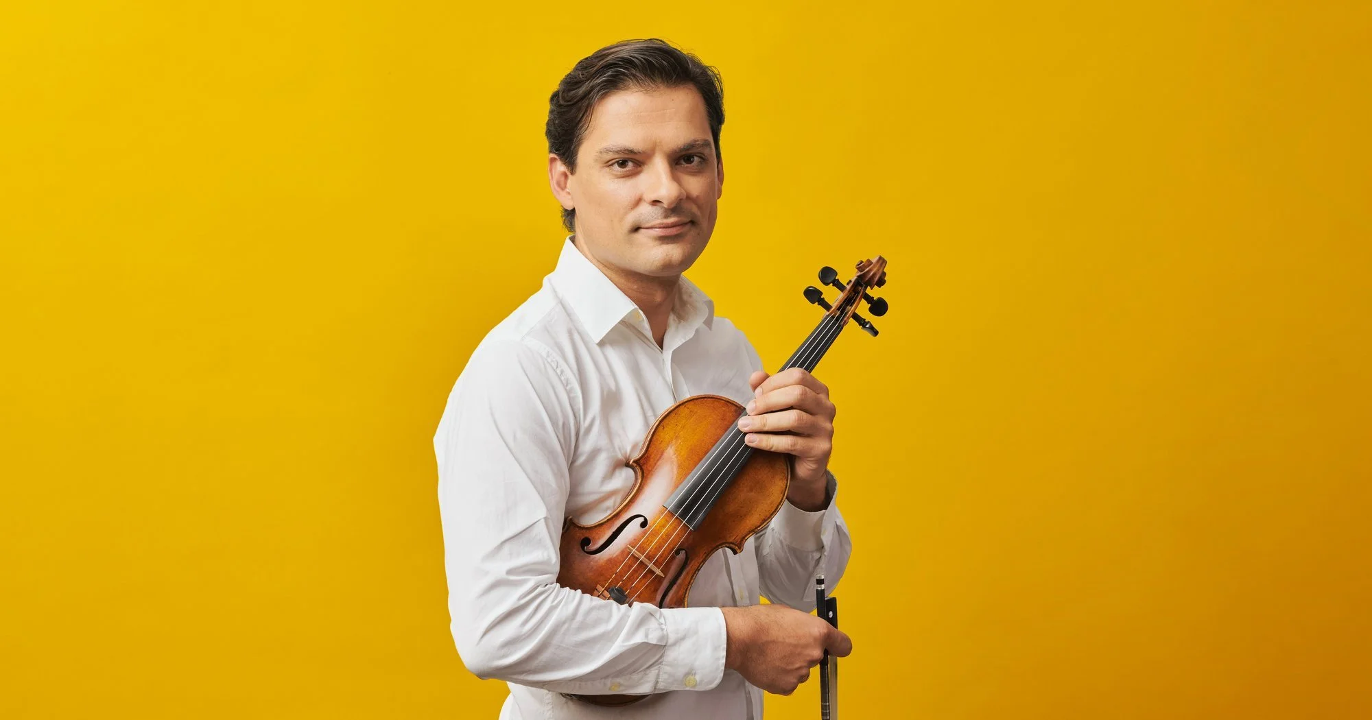 A man in a white shirt holding a violin and bow stands against a bright yellow background.