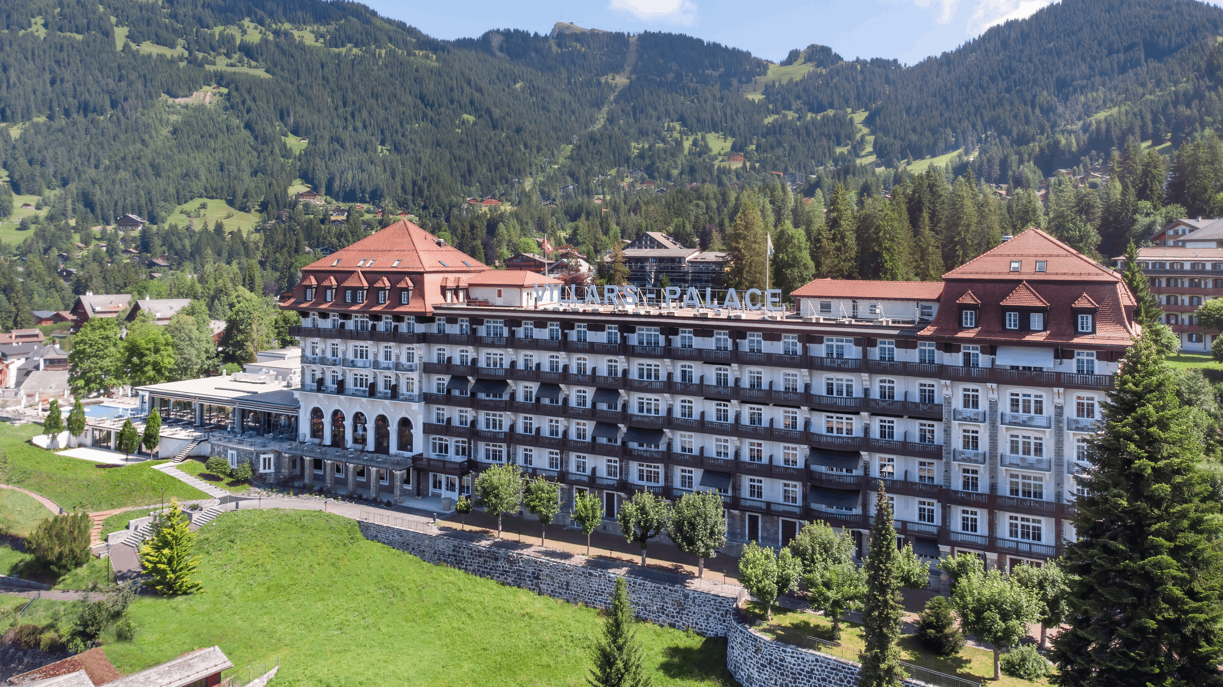 A large hotel building with multiple balconies and a sign that reads 'VILLARS PALACE' in a mountainous area with dense forests and green hills surrounding it.