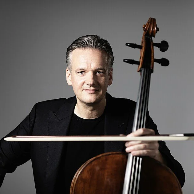 A man in a black jacket holding a cello and bow against a gray background.