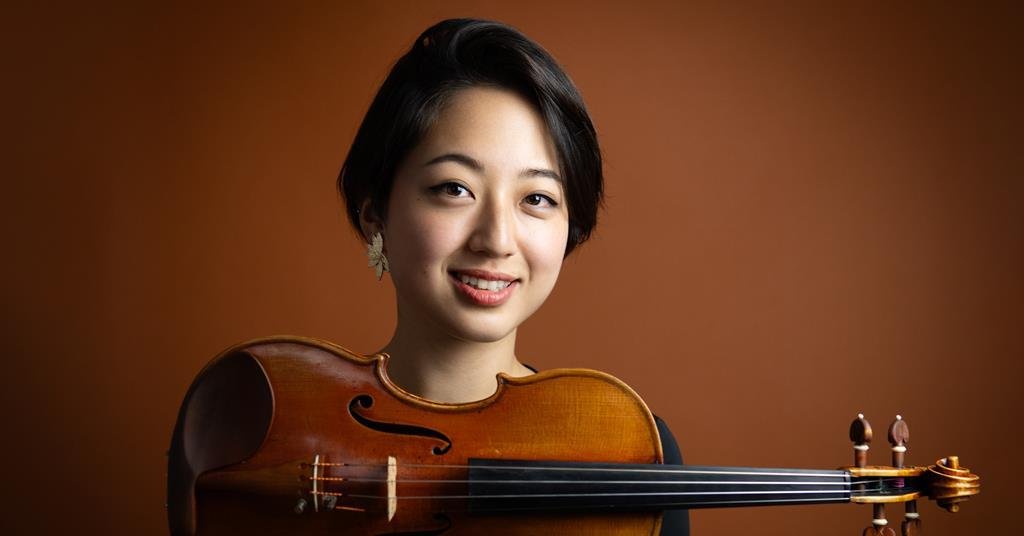 A young woman holding a violin in front of a brown background.