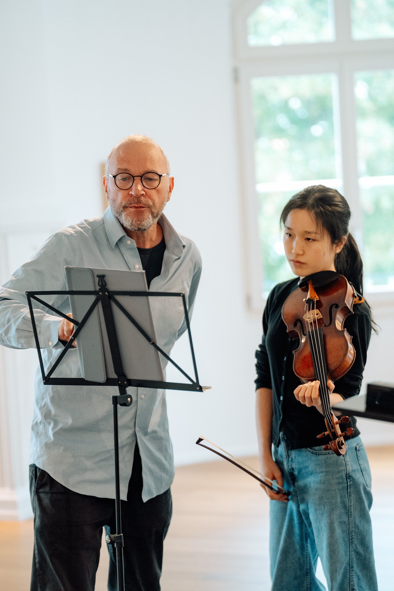 An older man with glasses and a beard holding sheet music, standing next to a young woman with a violin, both inside a room with large windows.