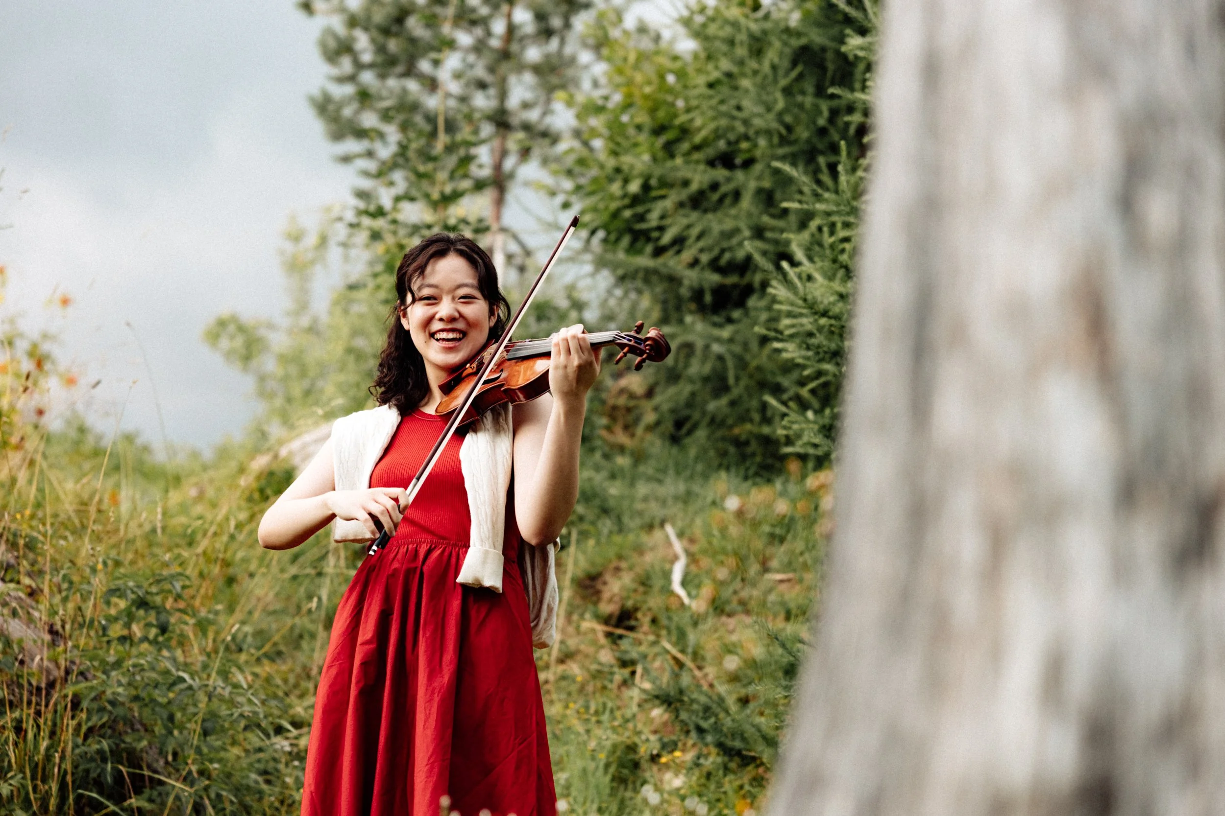 A young woman with short dark hair, wearing a red dress and white vest, playing a violin outdoors in a lush green natural setting.