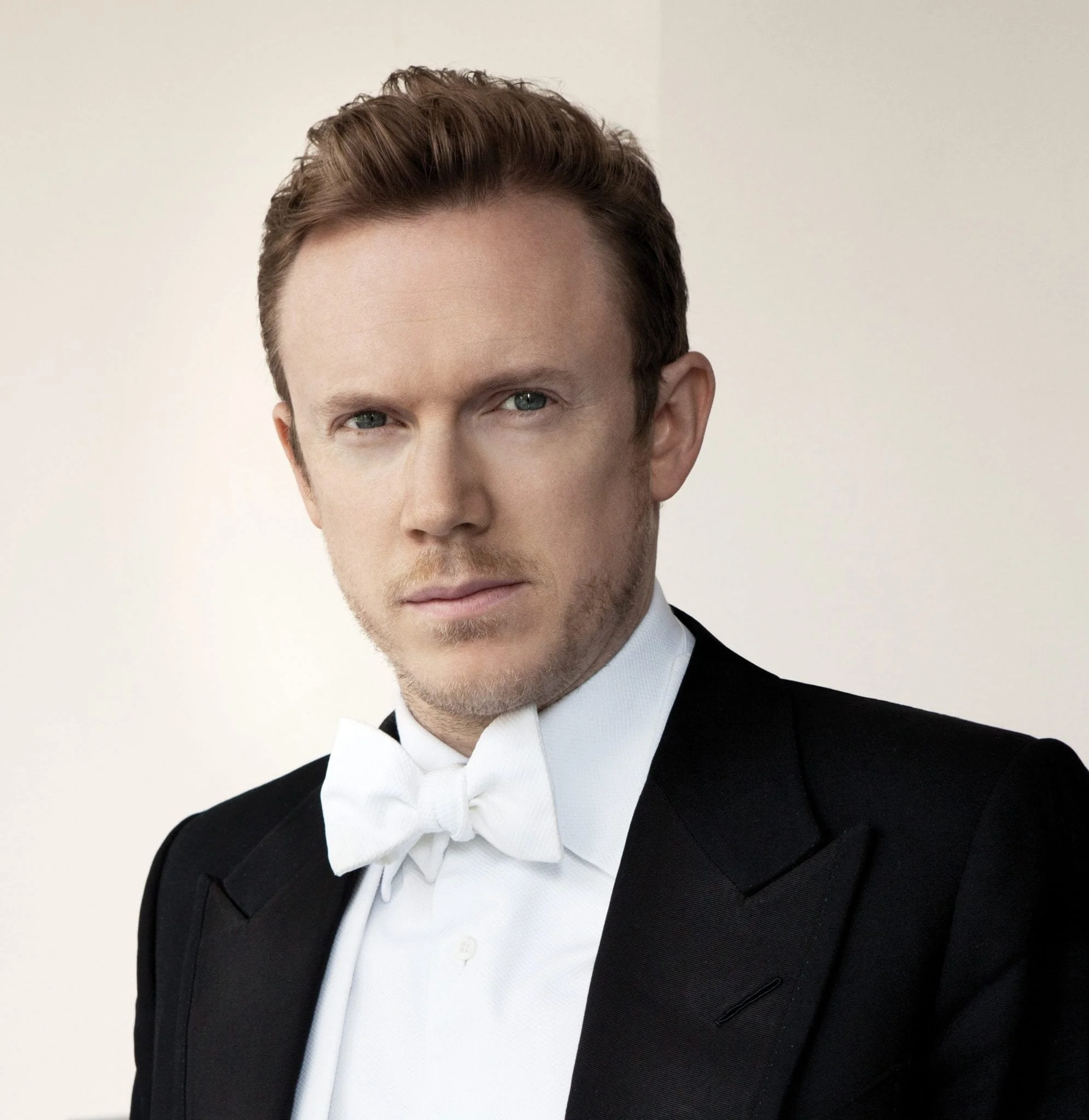 Close-up of a man wearing a black tuxedo with a satin lapel, white shirt, and white bow tie, against a plain light background.