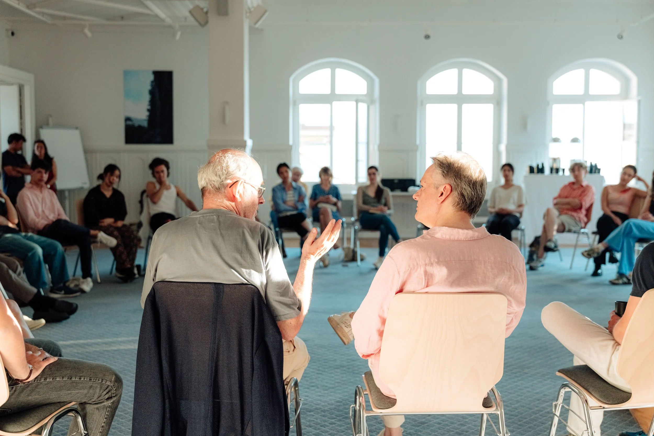 A group of people gathered in a spacious room with large windows, listening to a man with glasses who is speaking to a woman in a pink shirt. The participants are seated in chairs and appear to be engaged in a discussion or workshop.