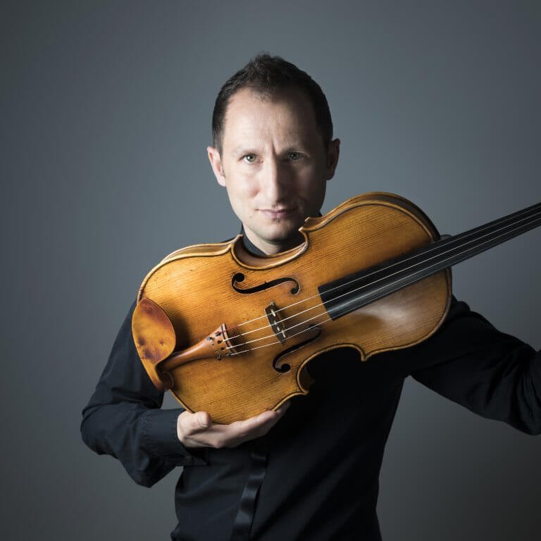 A man holding a violin across his chest, looking into the camera with a serious expression, against a gray background.