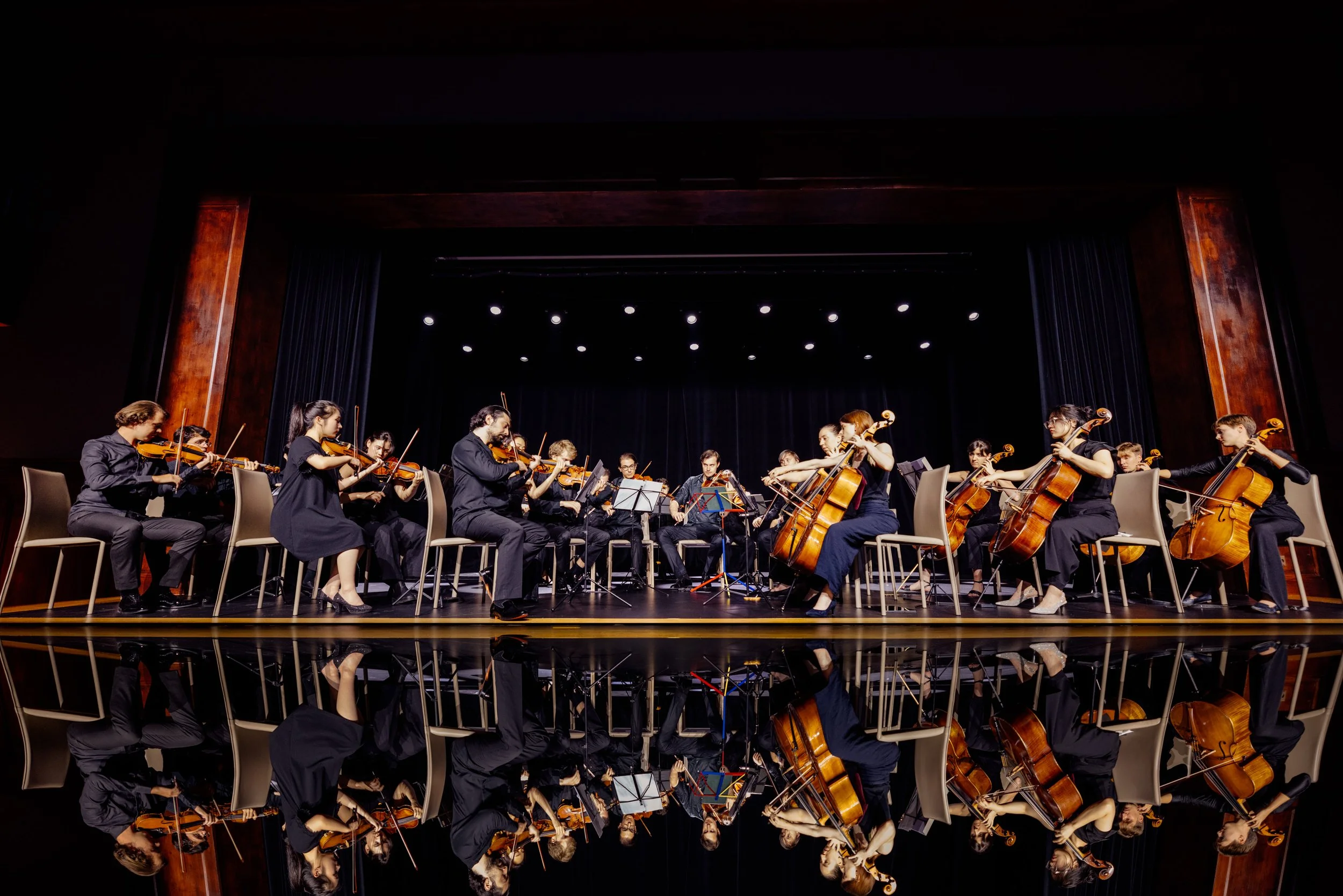 Orchestra performing on stage with string instruments, with a black curtain and stage lights in the background.