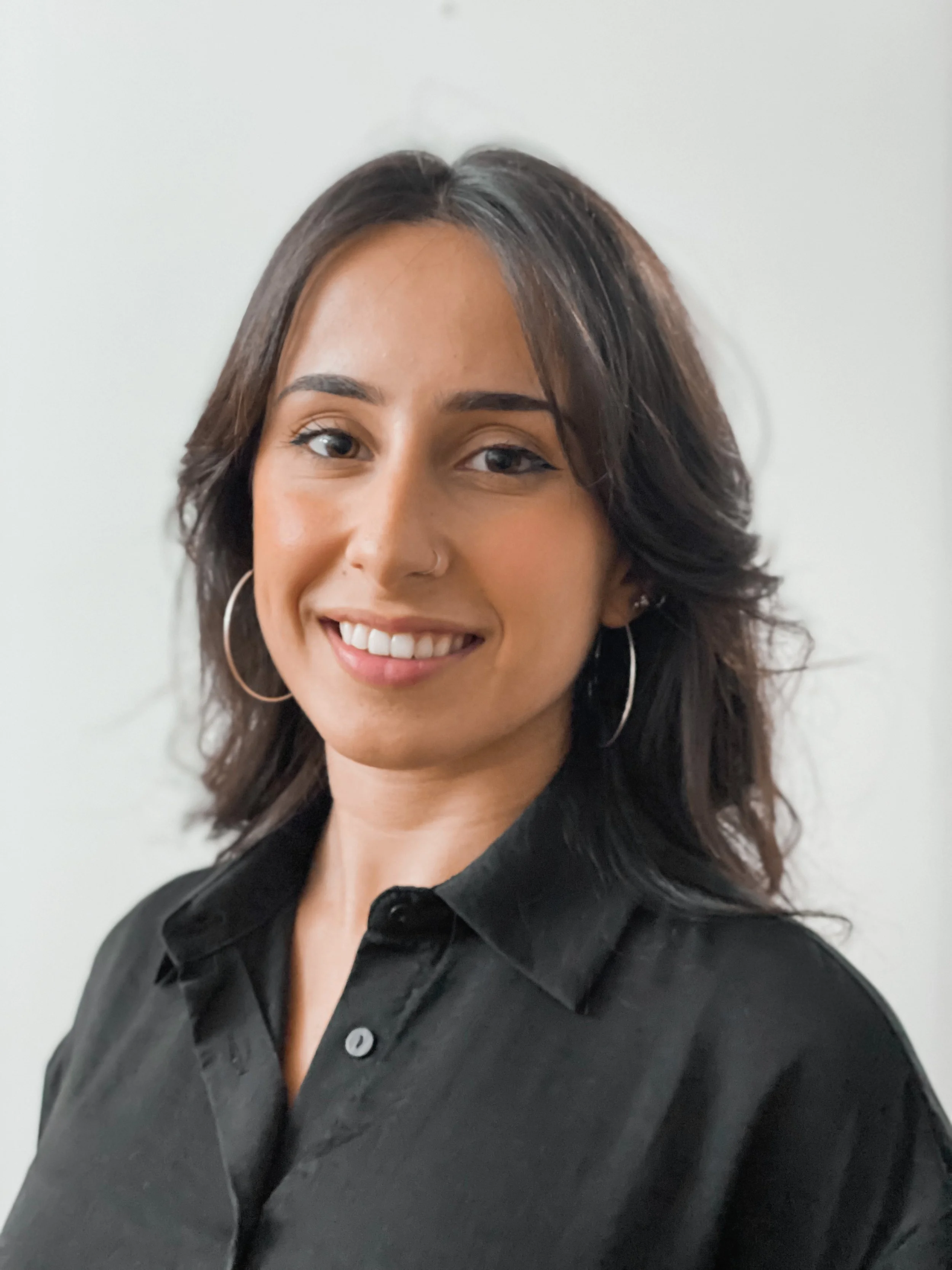 A woman with shoulder-length dark hair, wearing hoop earrings and a black shirt, smiling and looking at the camera, against a plain light-colored background.
