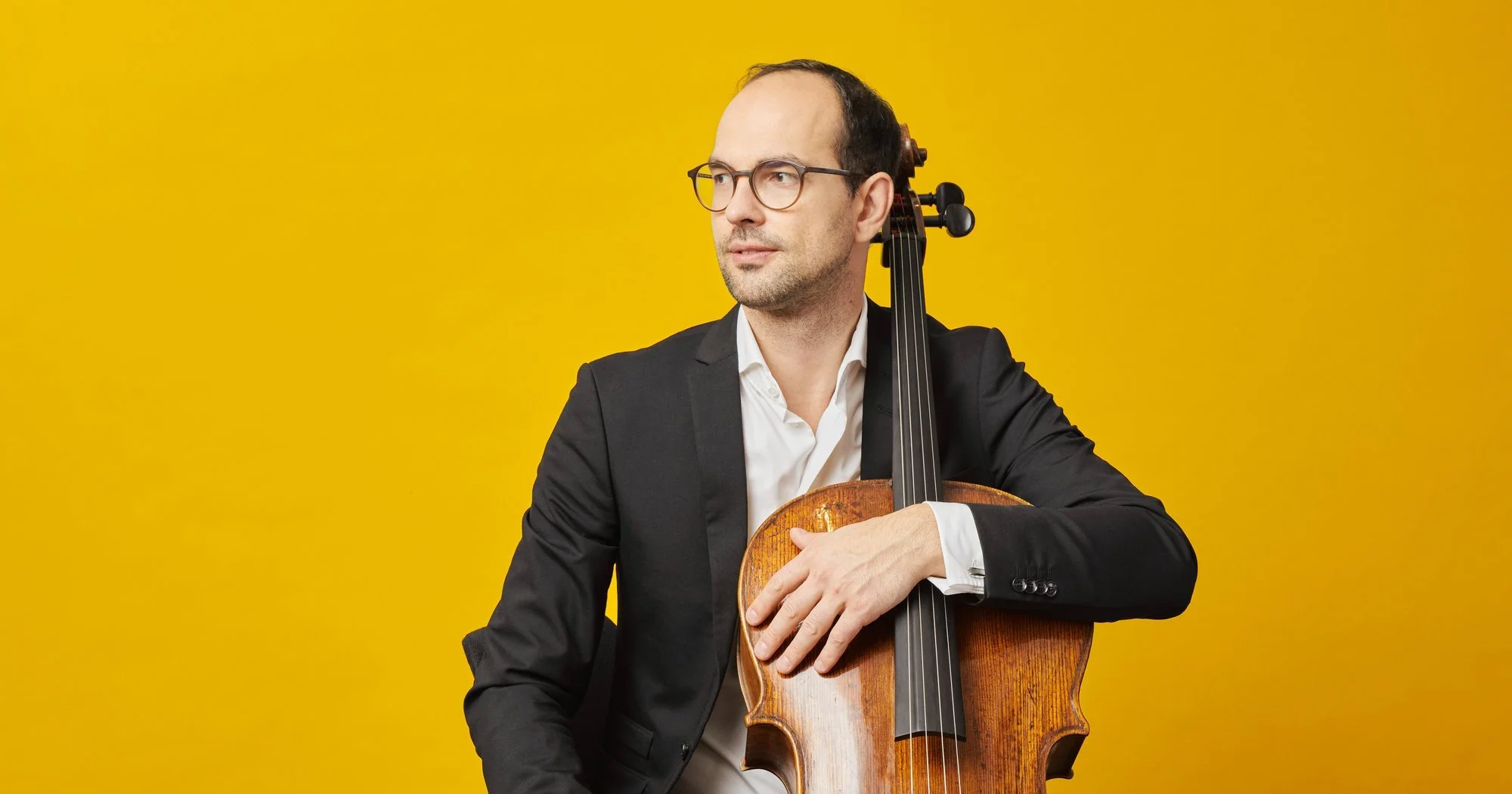 Man in a black suit and glasses holding a cello against a yellow background.