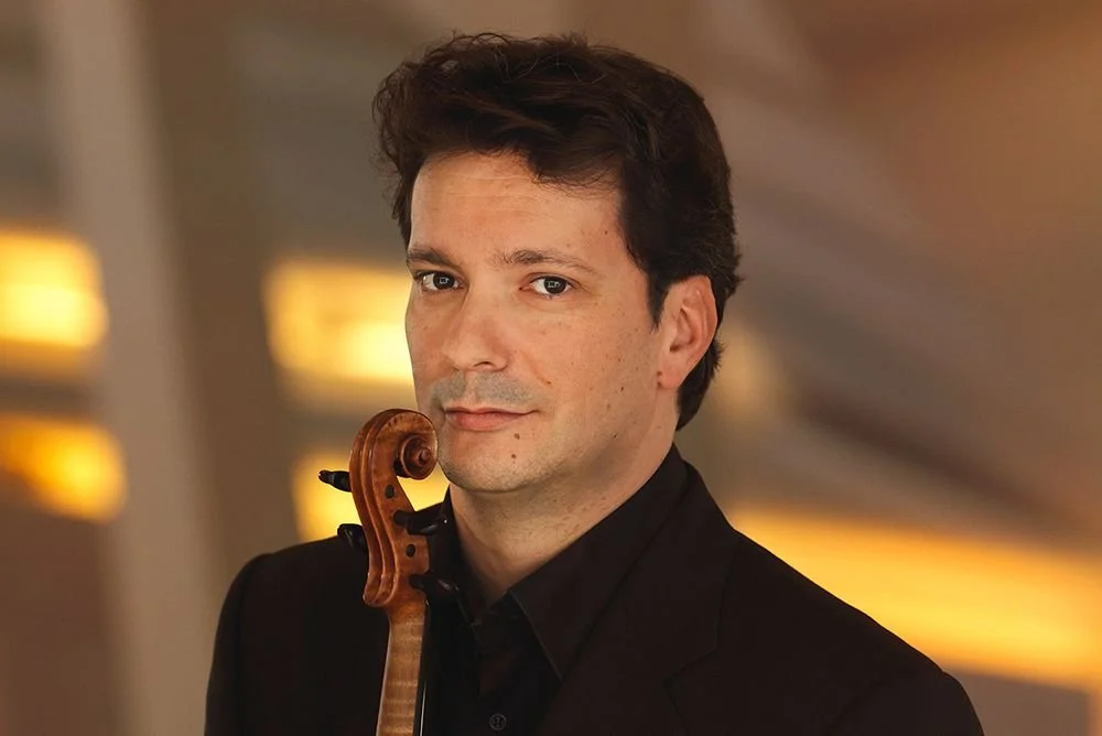 Man with dark hair and a black suit holding a cello upright, looking at the camera, indoor setting with warm yellow lighting.
