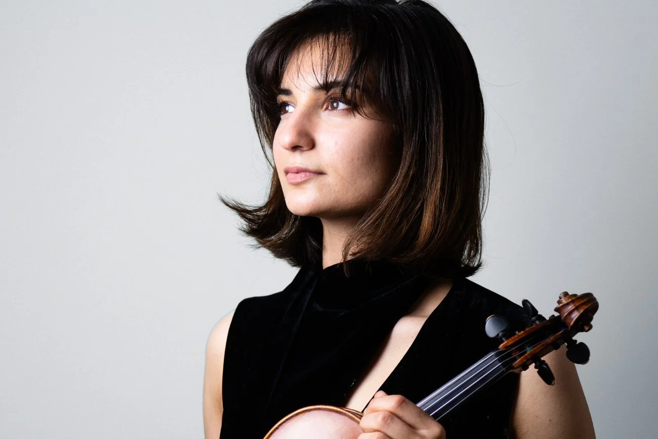 Woman holding a violin, looking to the left, with shoulder-length brown hair, wearing a sleeveless black top, against a plain white background.