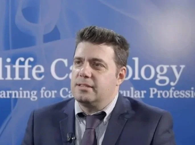 A man in a suit and tie speaking or being interviewed in front of a blue backdrop with the words 'life c...' visible.
