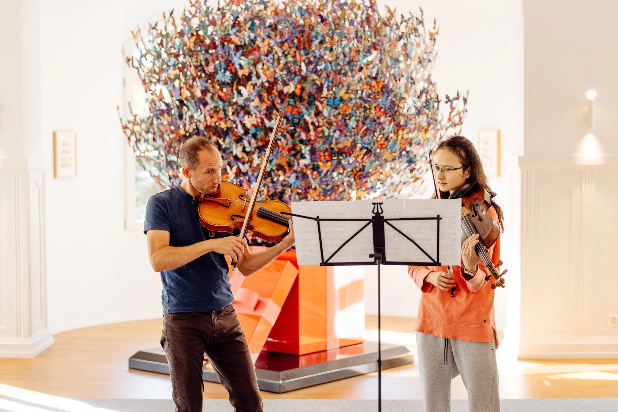 Two violinists practicing music with a colorful butterfly sculpture in the background.