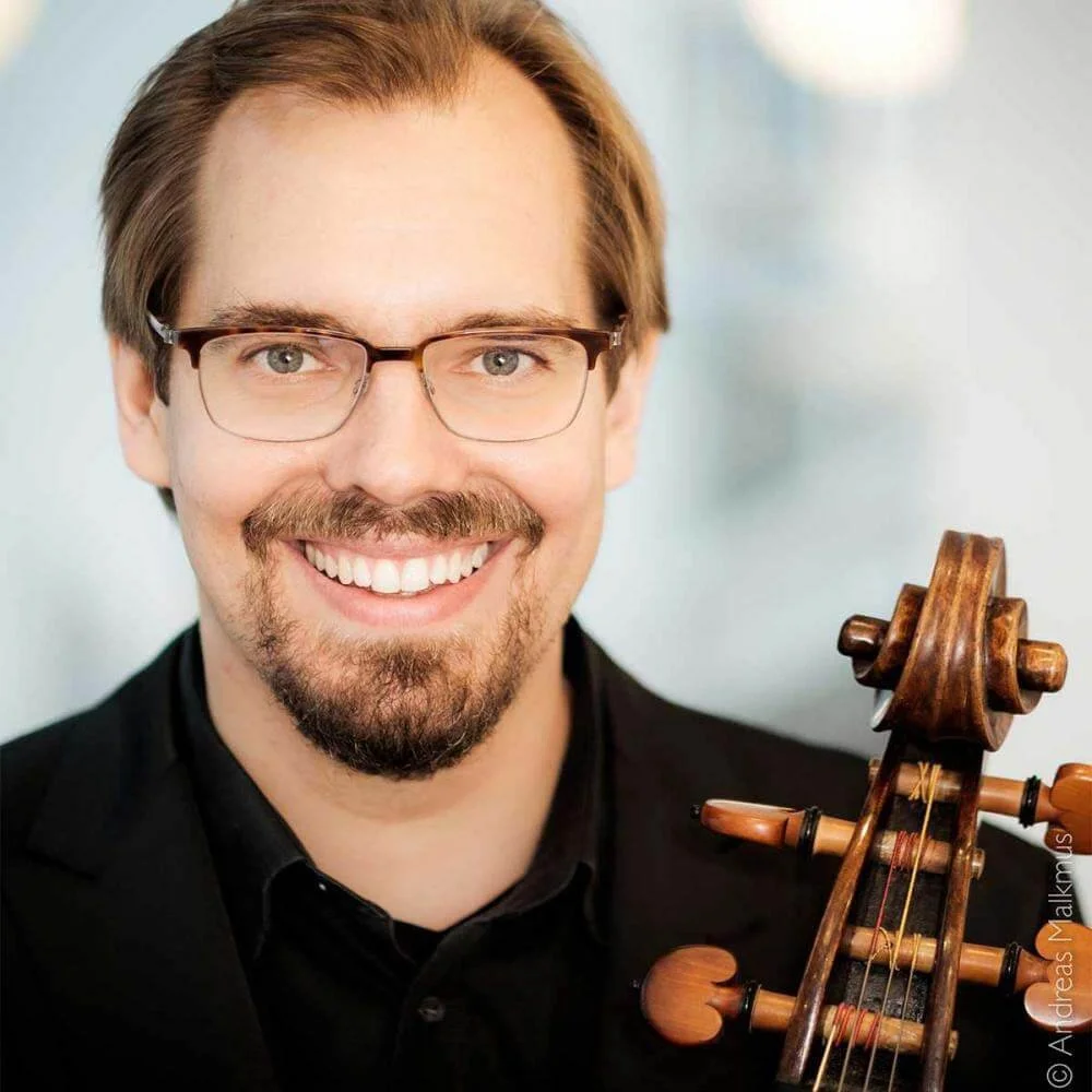A man with glasses, brown hair, and a beard smiling, holding a wooden violin.