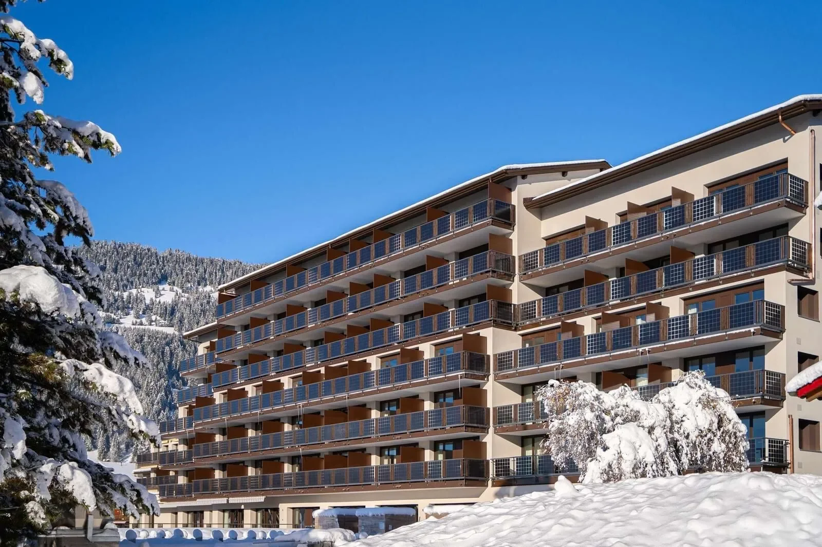 A large multi-story building with balconies, surrounded by snow-covered trees and mountains under a clear blue sky.