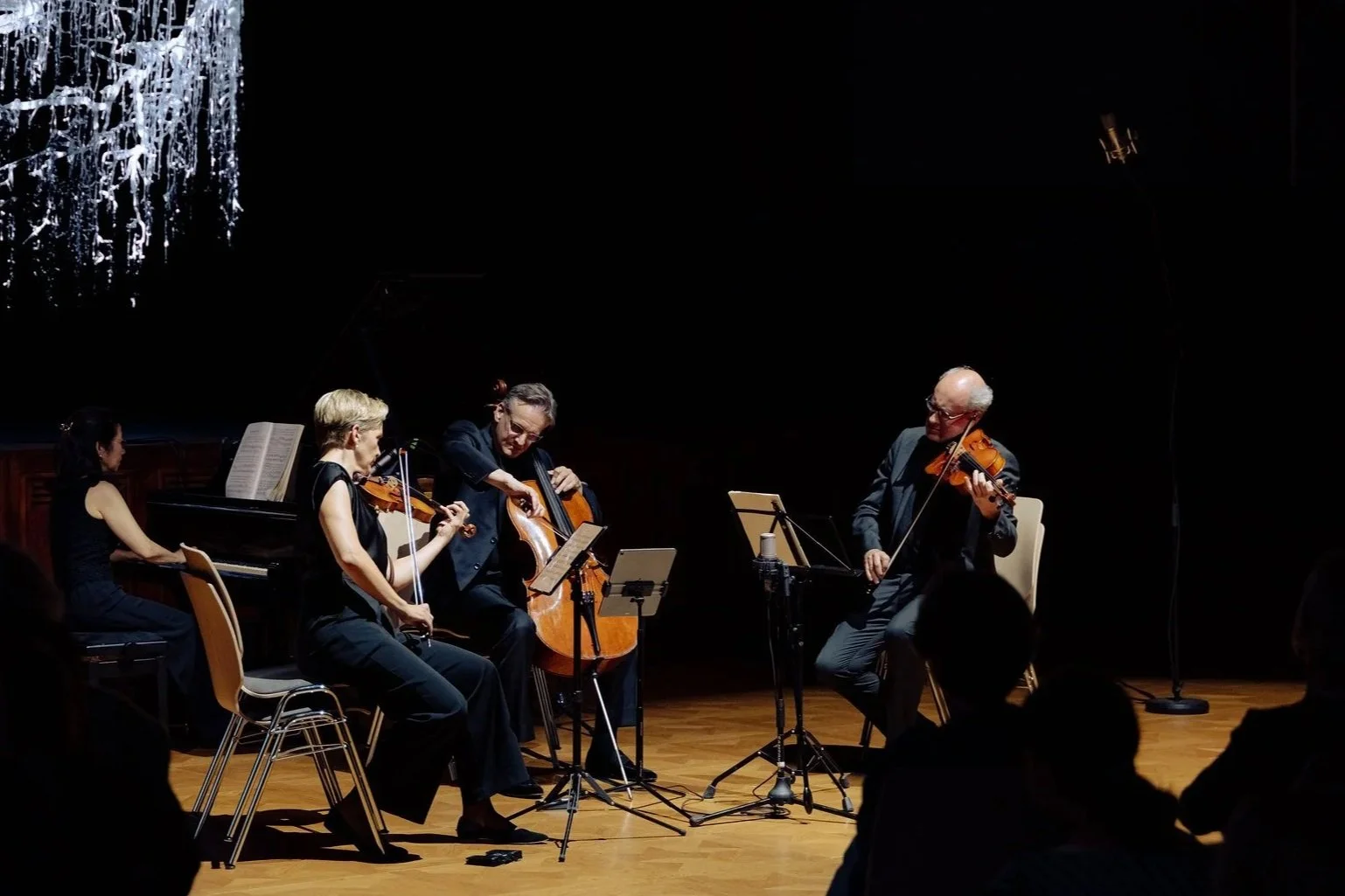 A group of four musicians performing on stage, playing piano, violin, and cello, with two audience members visible in the foreground, and a chandelier hanging from the ceiling.