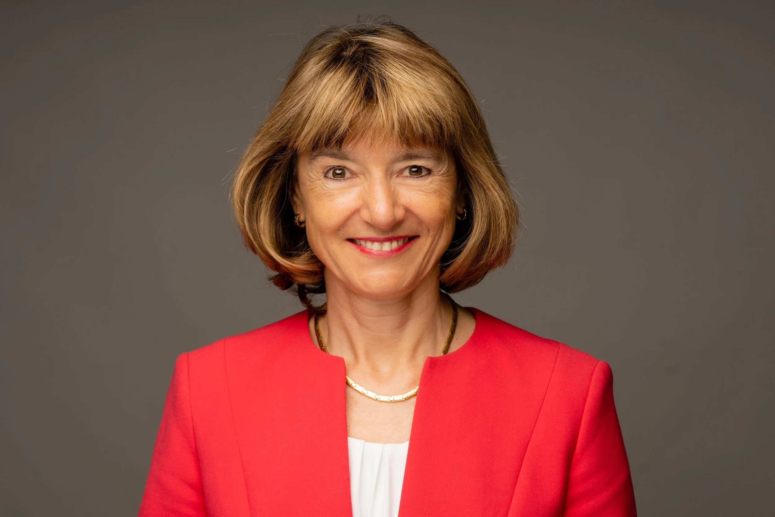 Portrait of a woman with light brown hair wearing a red blazer and a gold necklace, smiling against a gray background.