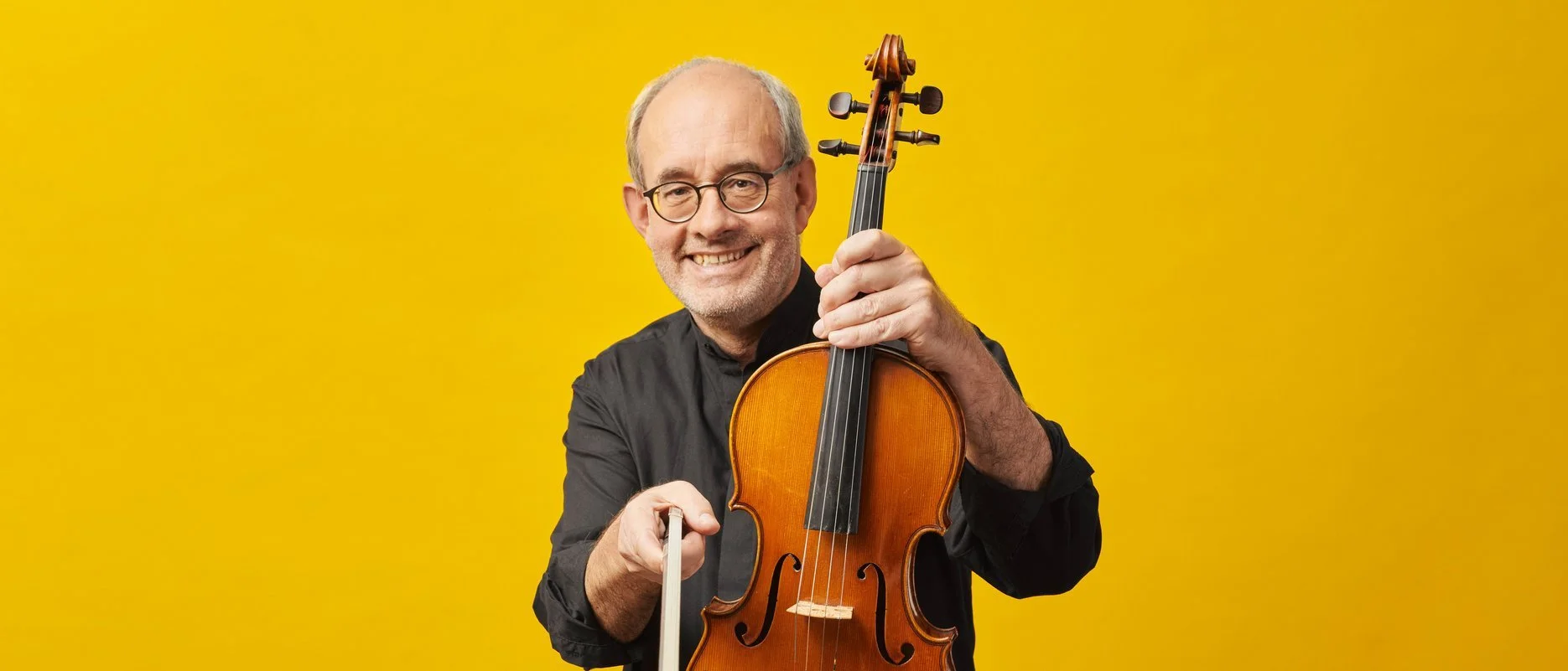 A smiling man with glasses holding a violin and bow against a yellow background.