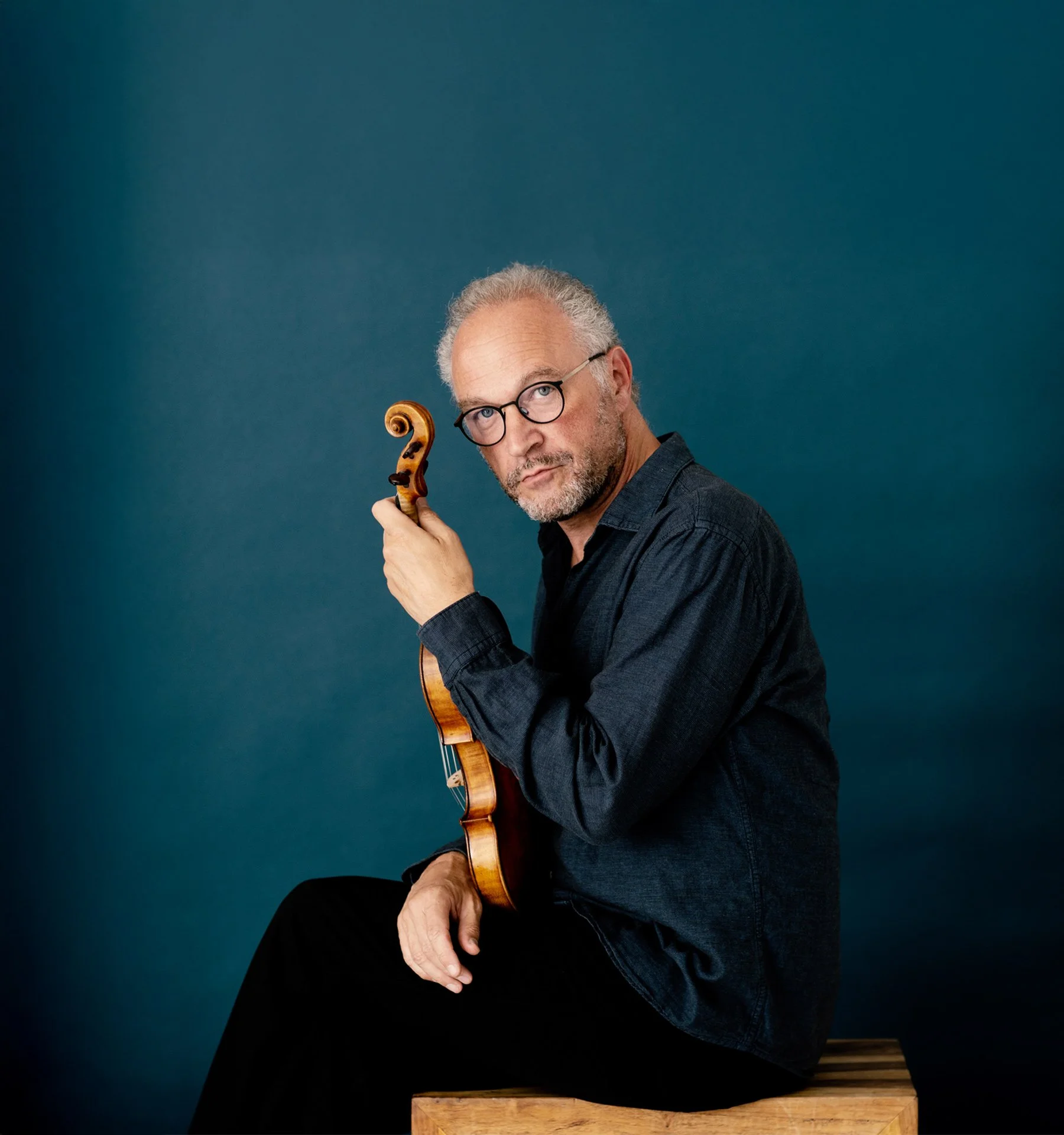 A man with gray hair and glasses holding a violin, sitting on a wooden chair against a blue background.