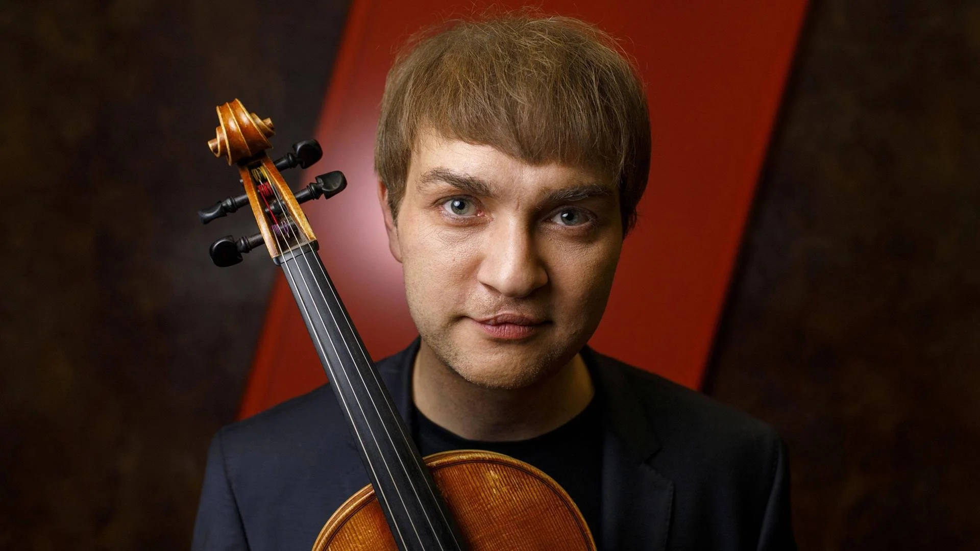 A man with light brown hair and blue eyes poses with a violin resting on his shoulder, against a dark background with red and black accents.