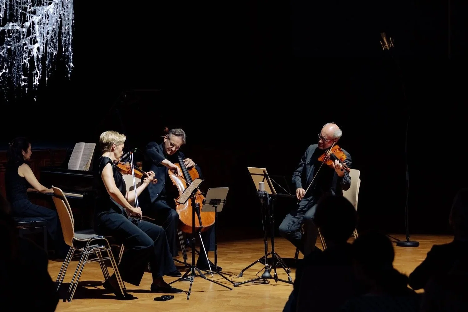 A group of five musicians performing on a dark stage, with a grand piano to the left and standing microphones nearby. They are playing string instruments: two violins, a cello, and a viola.