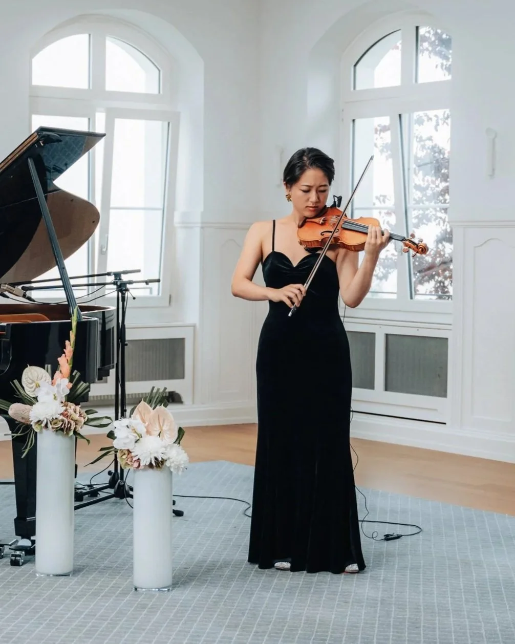 A woman in a long black dress playing the violin in a bright room with large windows, a grand piano, and floral arrangements.
