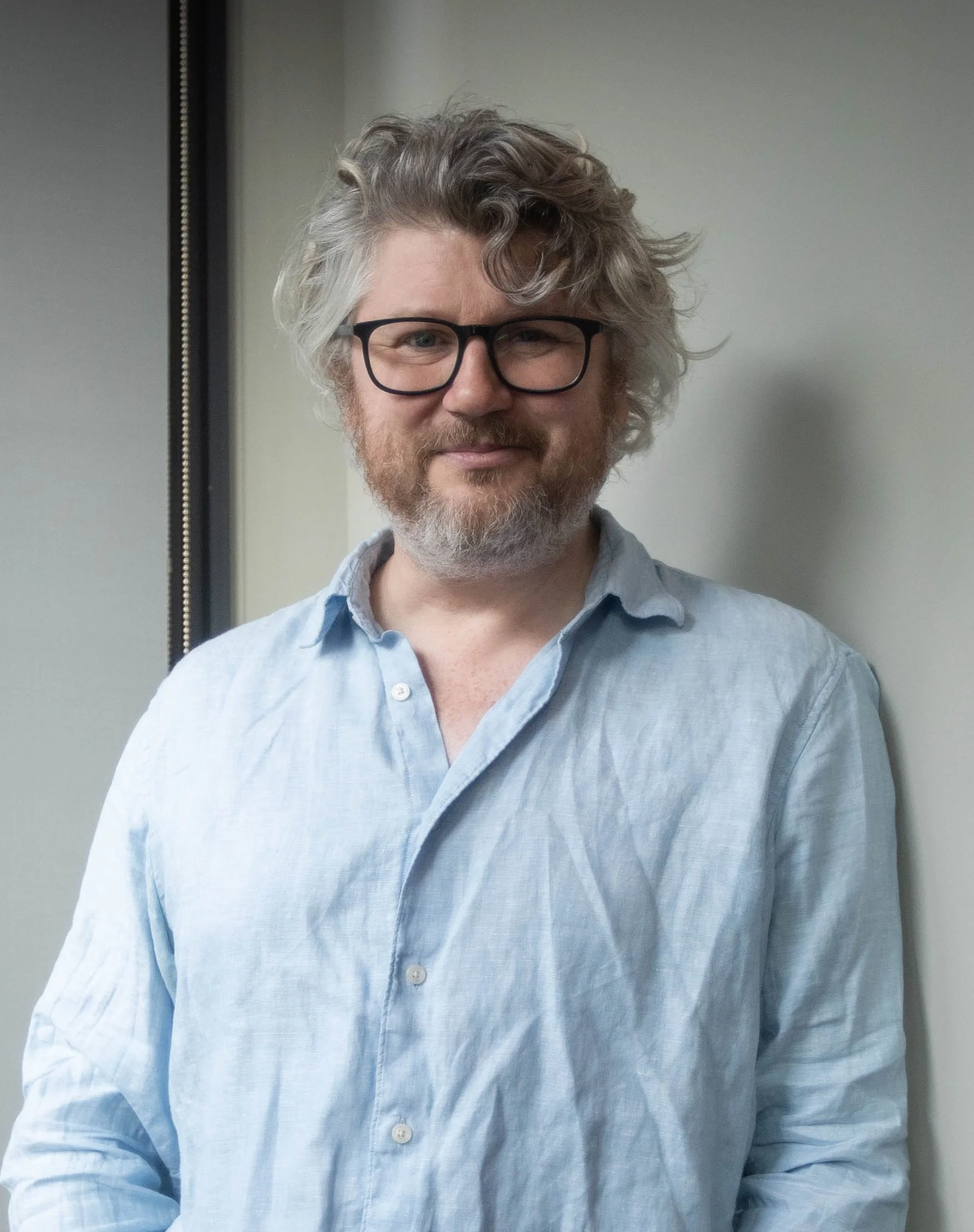 A man with curly hair, wearing glasses and a light blue shirt, standing indoors against a plain wall.