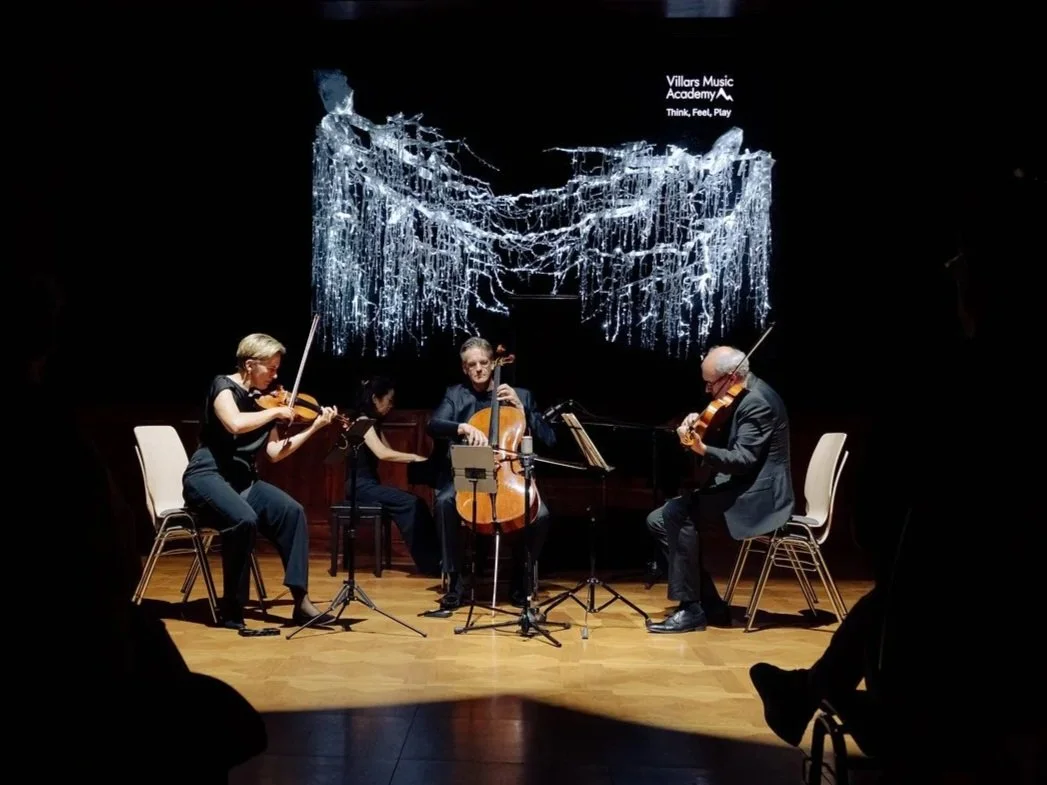 Four musicians playing string instruments on stage with a dark background, and a projection that says 'Villars Music Academy' and 'Think, Feel, Play'.