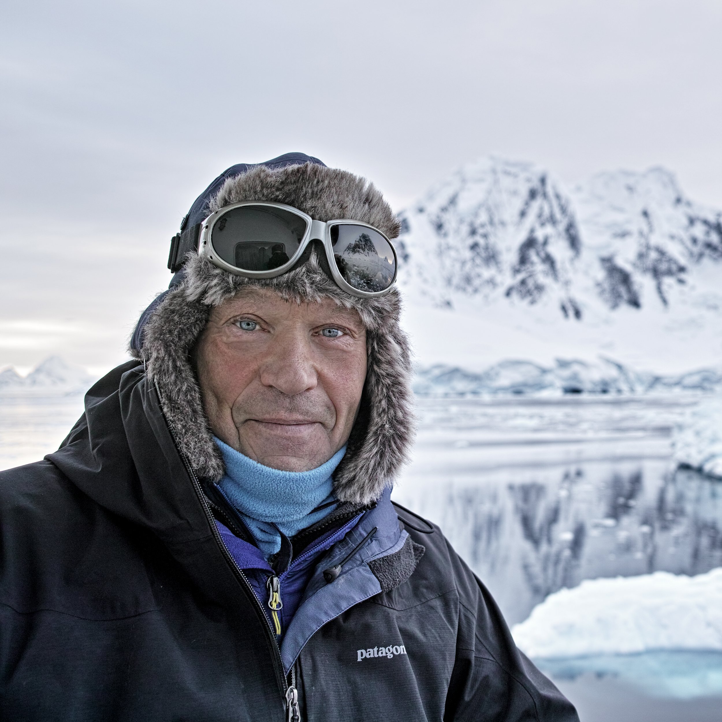 Close-up of a man wearing a warm hat and ski goggles, with snow-covered mountains in the background.