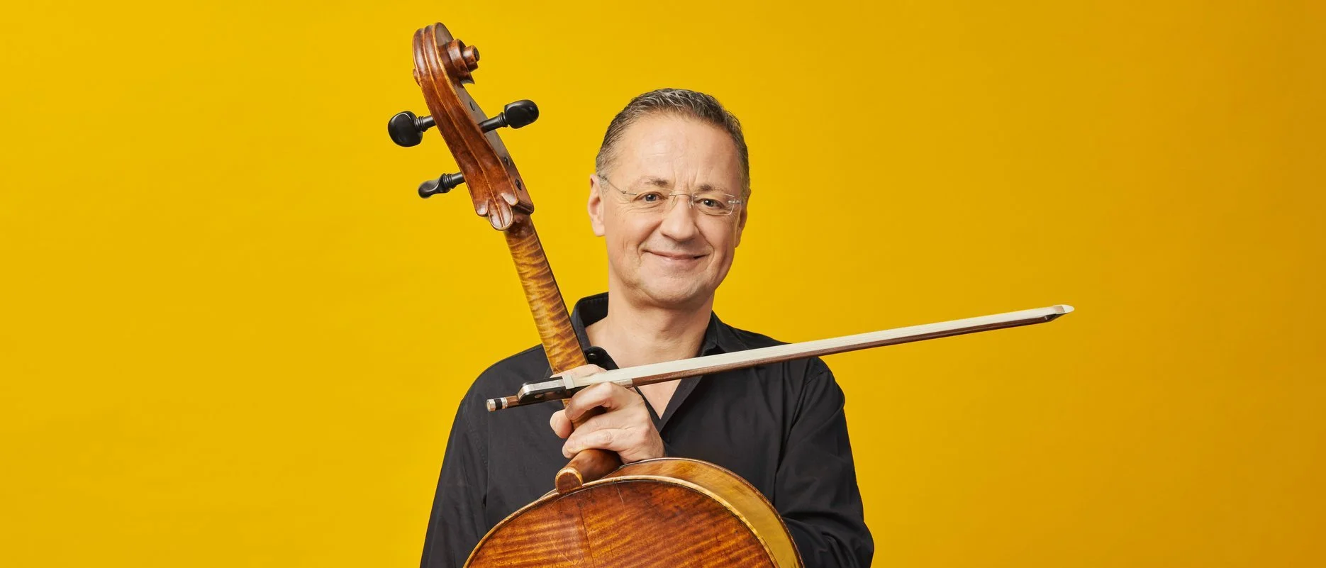 A man with glasses in a black shirt smiling and holding a violin and bow in front of a solid yellow background.